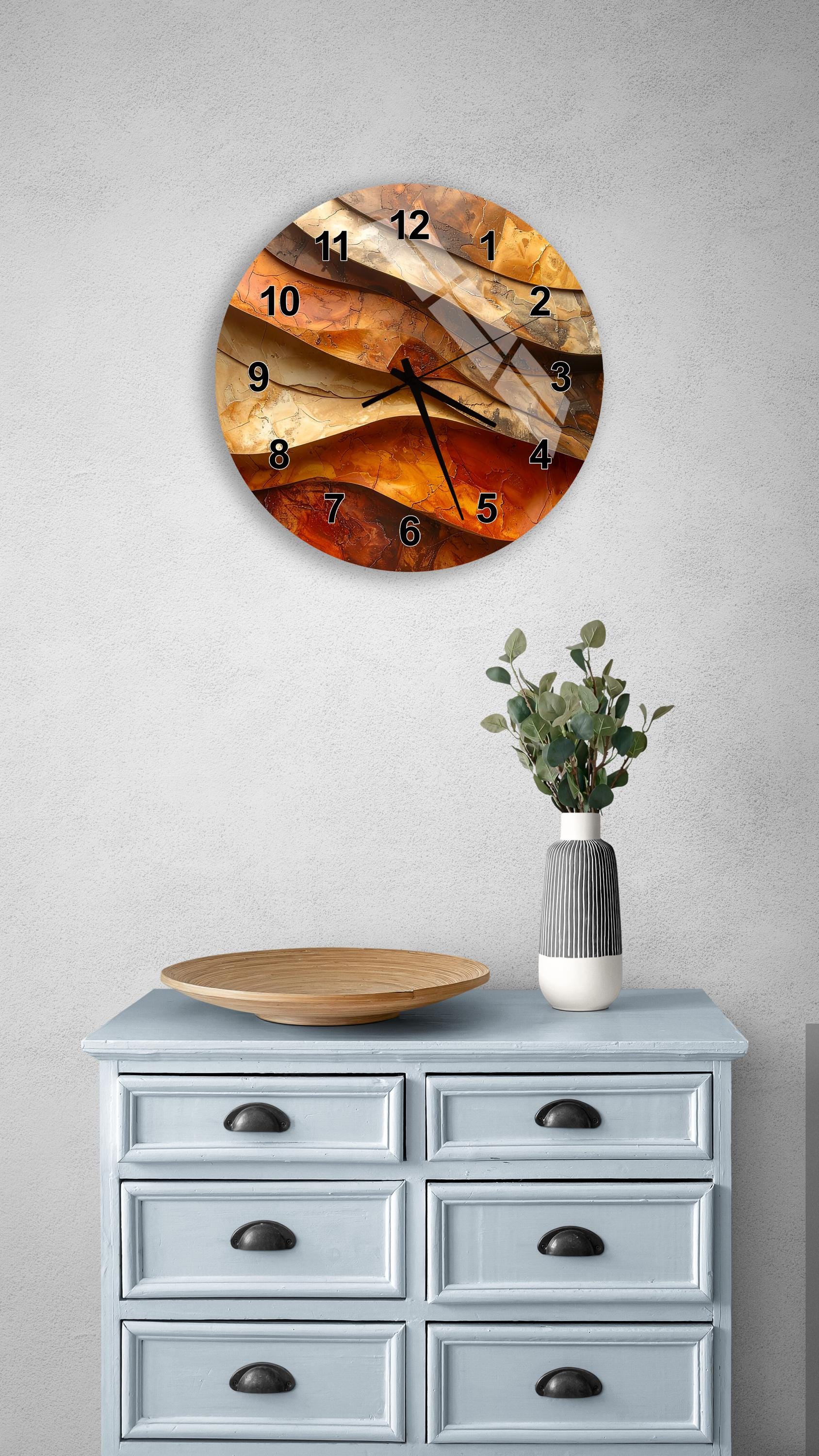 a wall clock with a unique abstract design, mounted on a wall above a light blue dresser with six drawers. The dresser also has a vase with greenery on it, and there is a wooden plate on top of the dresser.