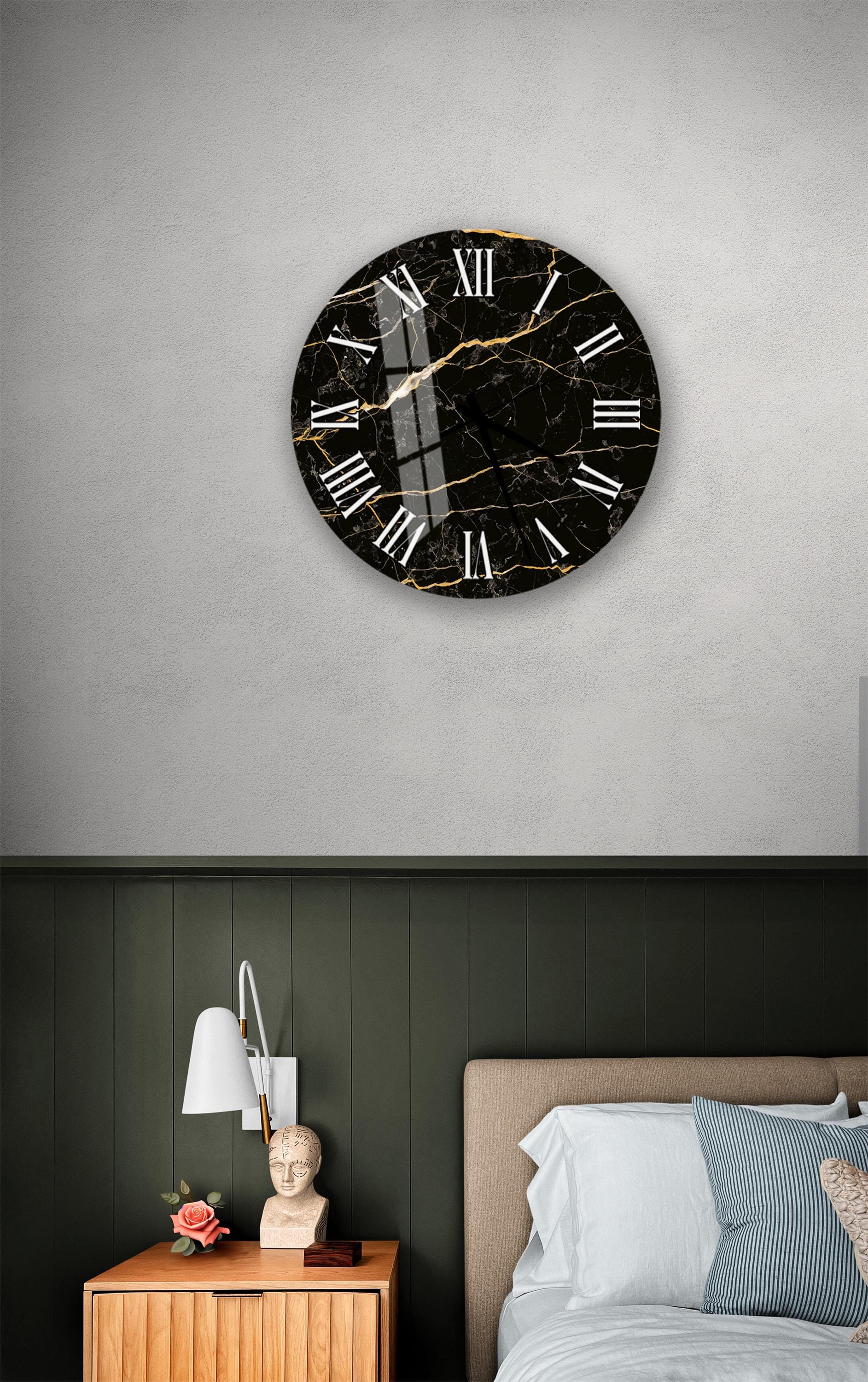 a bedroom setting with a large black and gold clock mounted on the wall above a bed. The clock has a marble-like finish and Roman numerals. The room has a green wall and a wooden nightstand with a lamp and a vase of flowers.