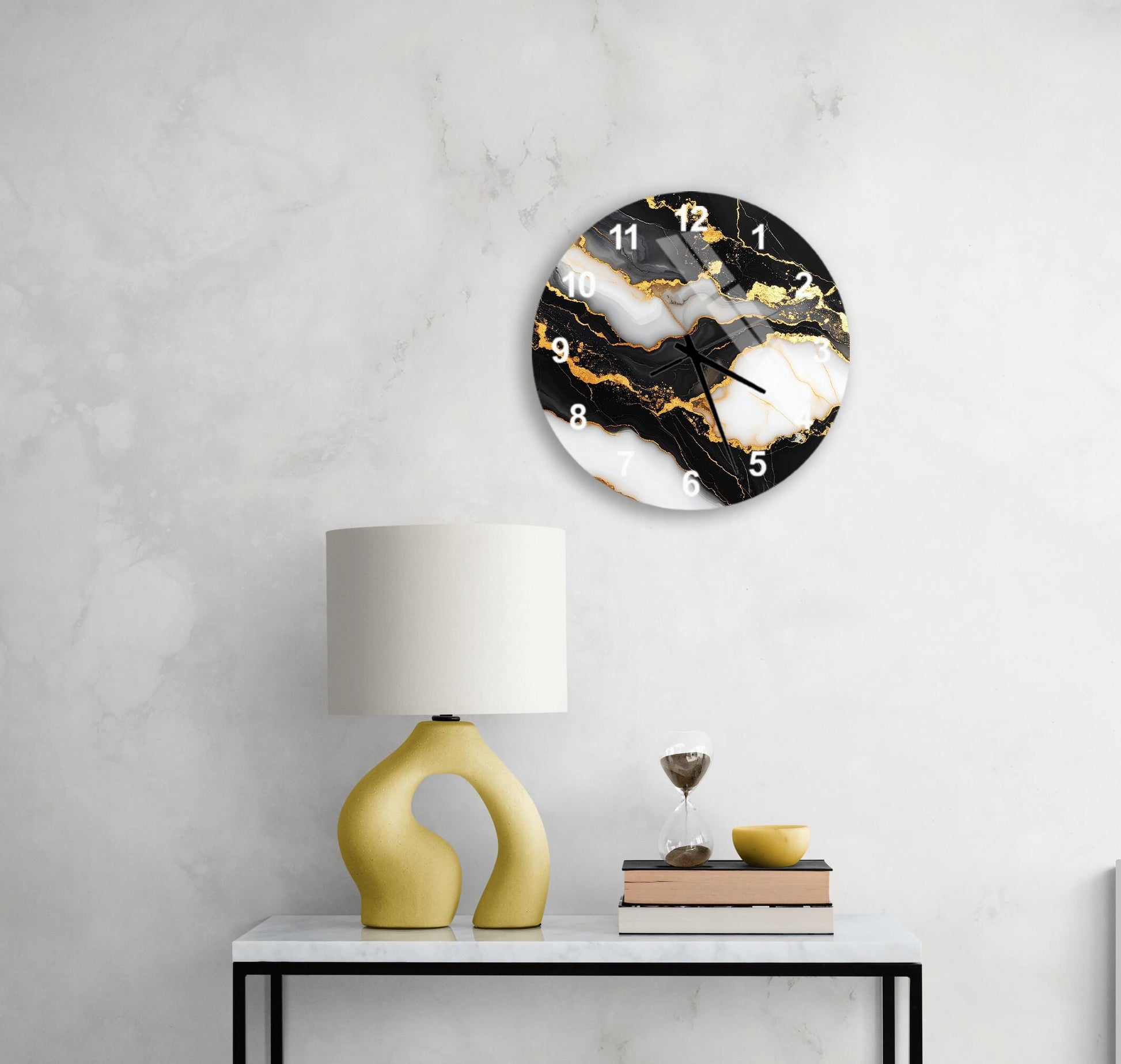 a modern black and white clock with gold accents, mounted on a white wall above a white table with a yellow lamp and a stack of books.