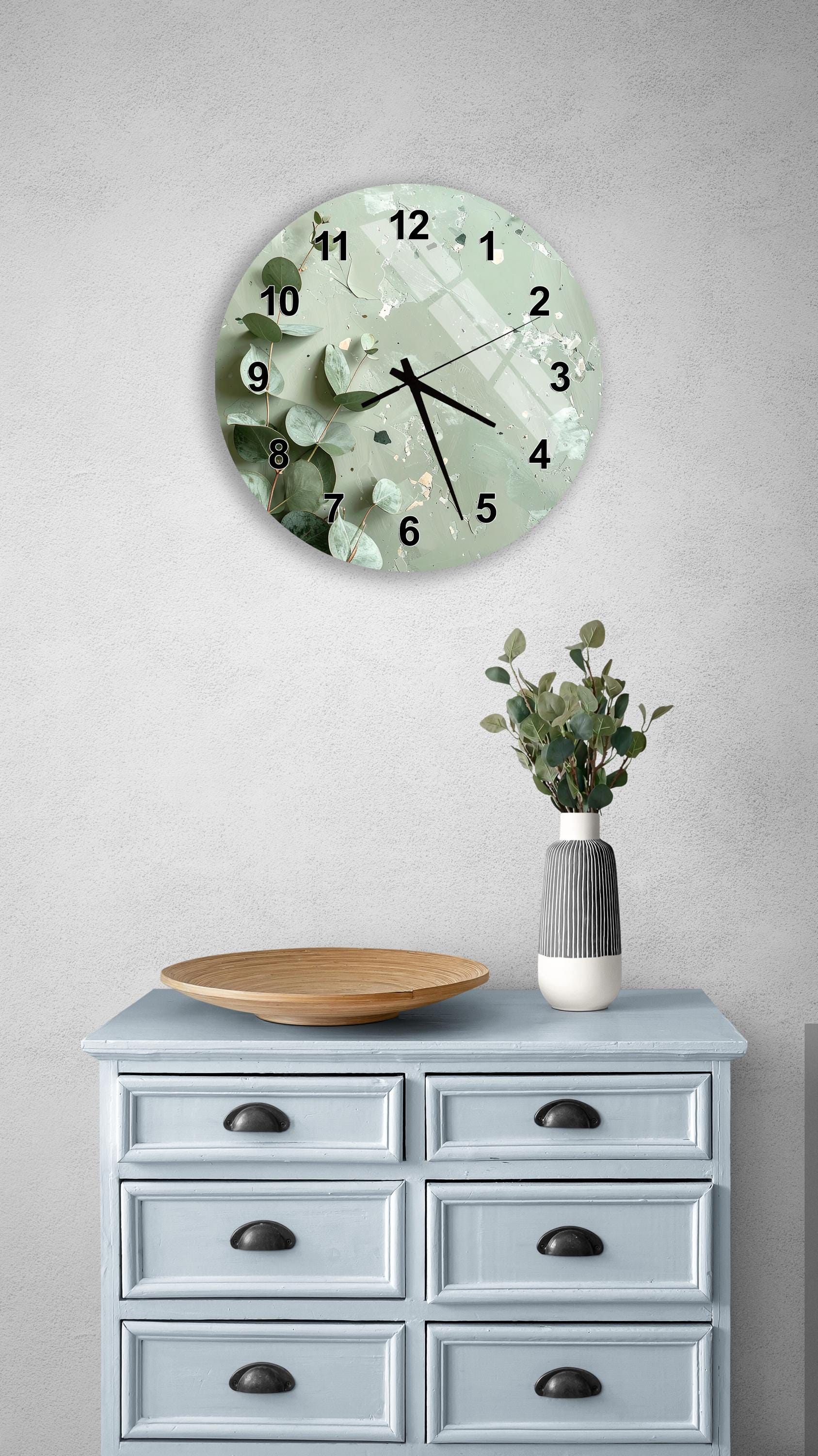 a wall clock with a green and white floral design, mounted on a wall above a light blue wooden dresser with six drawers. The dresser also has a vase with greenery on it, and a wooden plate on top.