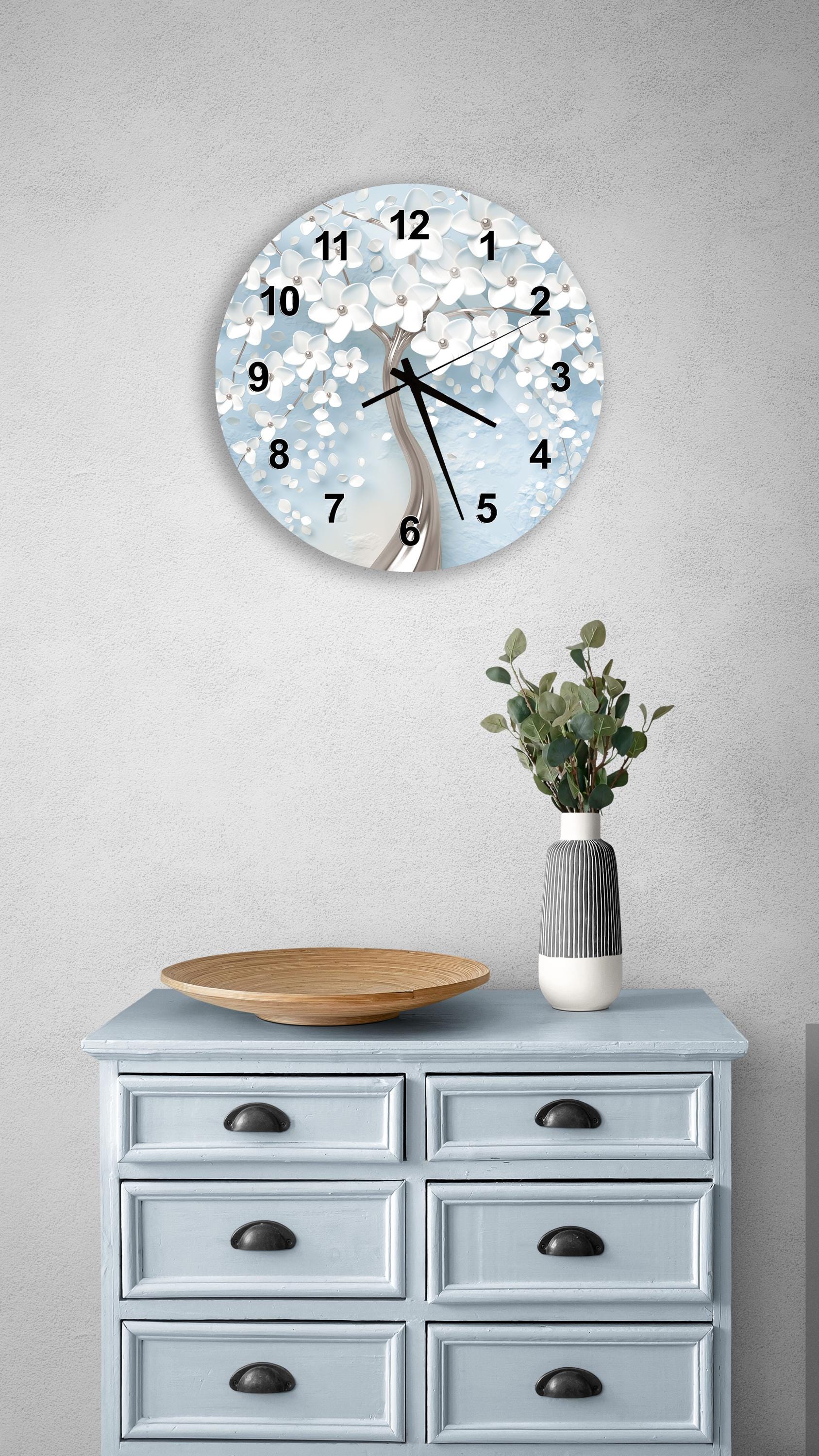 a wall clock with a white floral design, a wooden bowl, and a vase with greenery on a light blue dresser.
