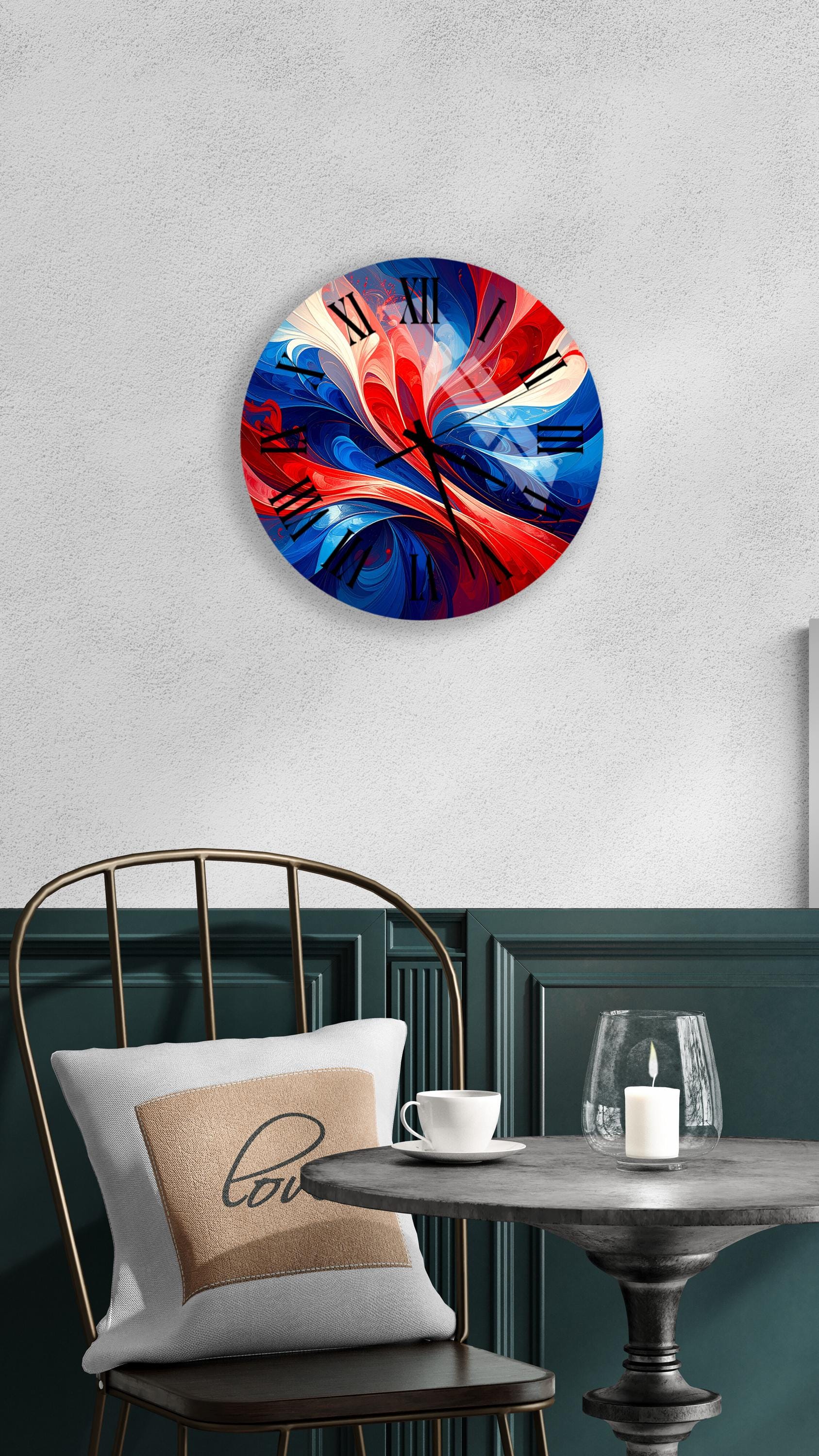 a round wall clock with a vibrant, abstract design in red, white, and blue colors. The clock is mounted on a wall above a dining table with a white tablecloth. A white cup and a candle are placed on the table, creating a cozy and inviting atmosphere.