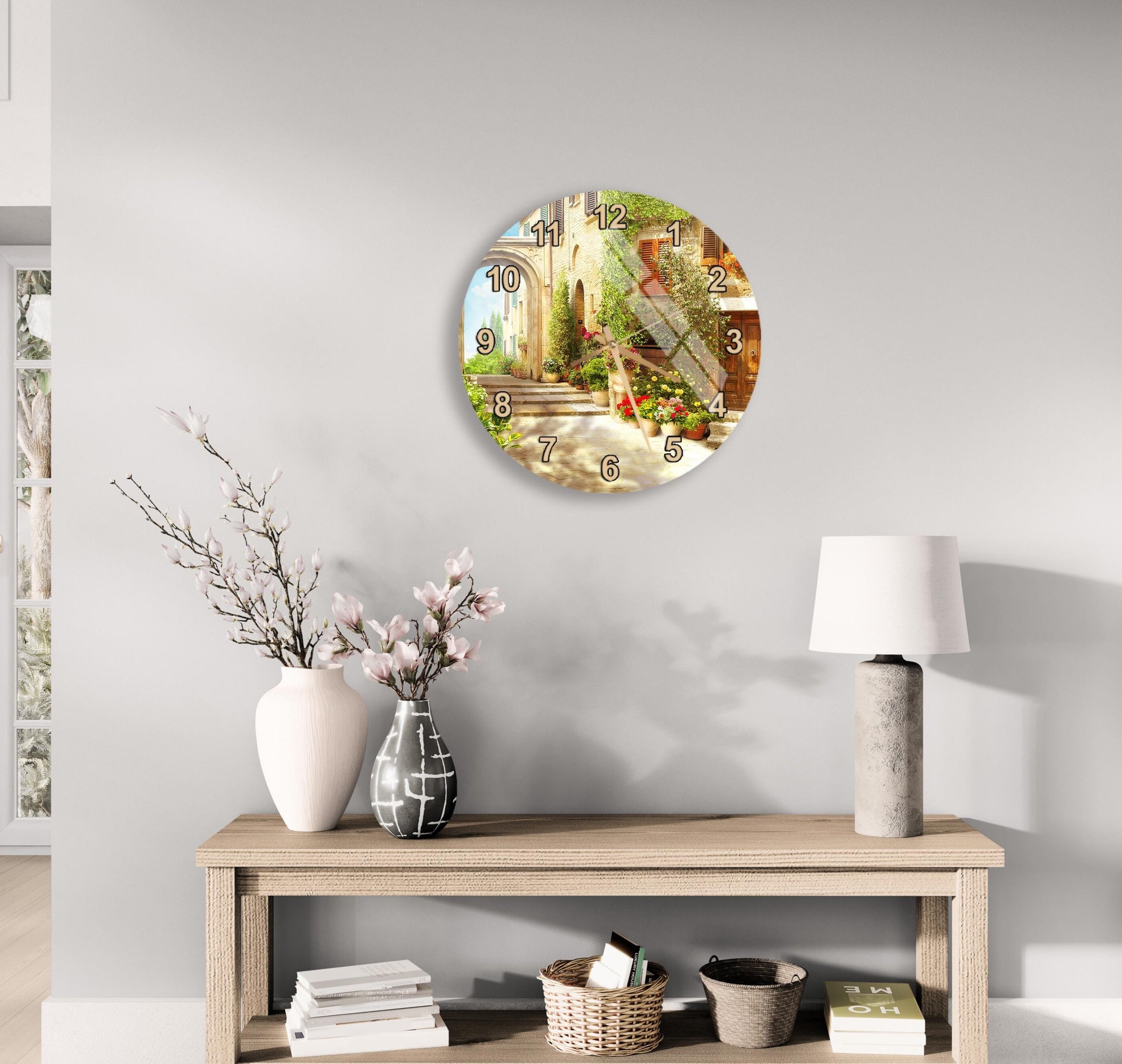 a wall clock with a scenic outdoor view, a wooden table with a lamp, a vase with flowers, and some books on a shelf.