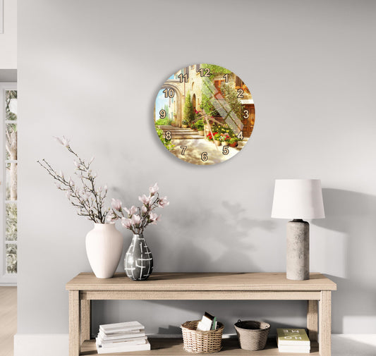 a wall clock with a scenic outdoor view, a wooden table with a lamp, a vase with flowers, and some books on a shelf.