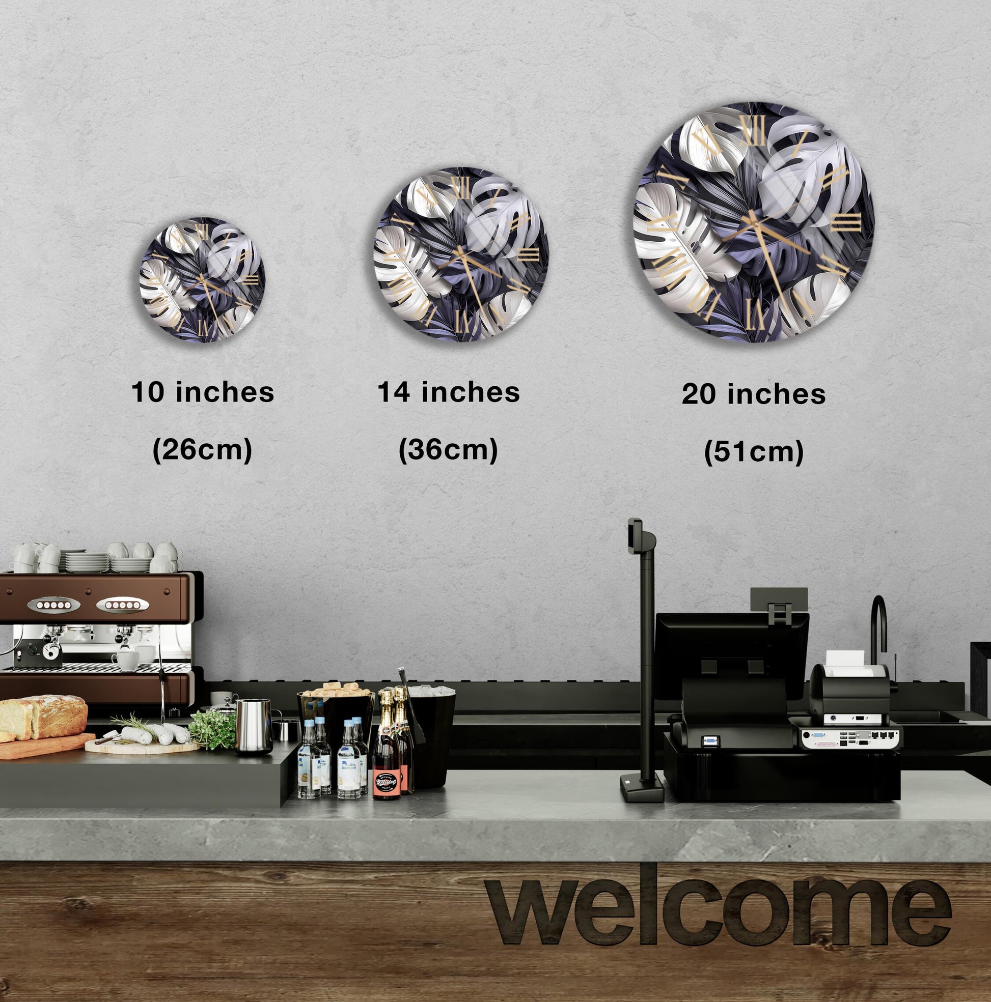 A wall clock with a tropical leaf pattern is displayed on a wall, accompanied by three different sizes of the clock, ranging from 10 inches to 20 inches. Below the clock, there is a counter with various items, including bottles and a sandwich. The word "welcome" is written on the counter.