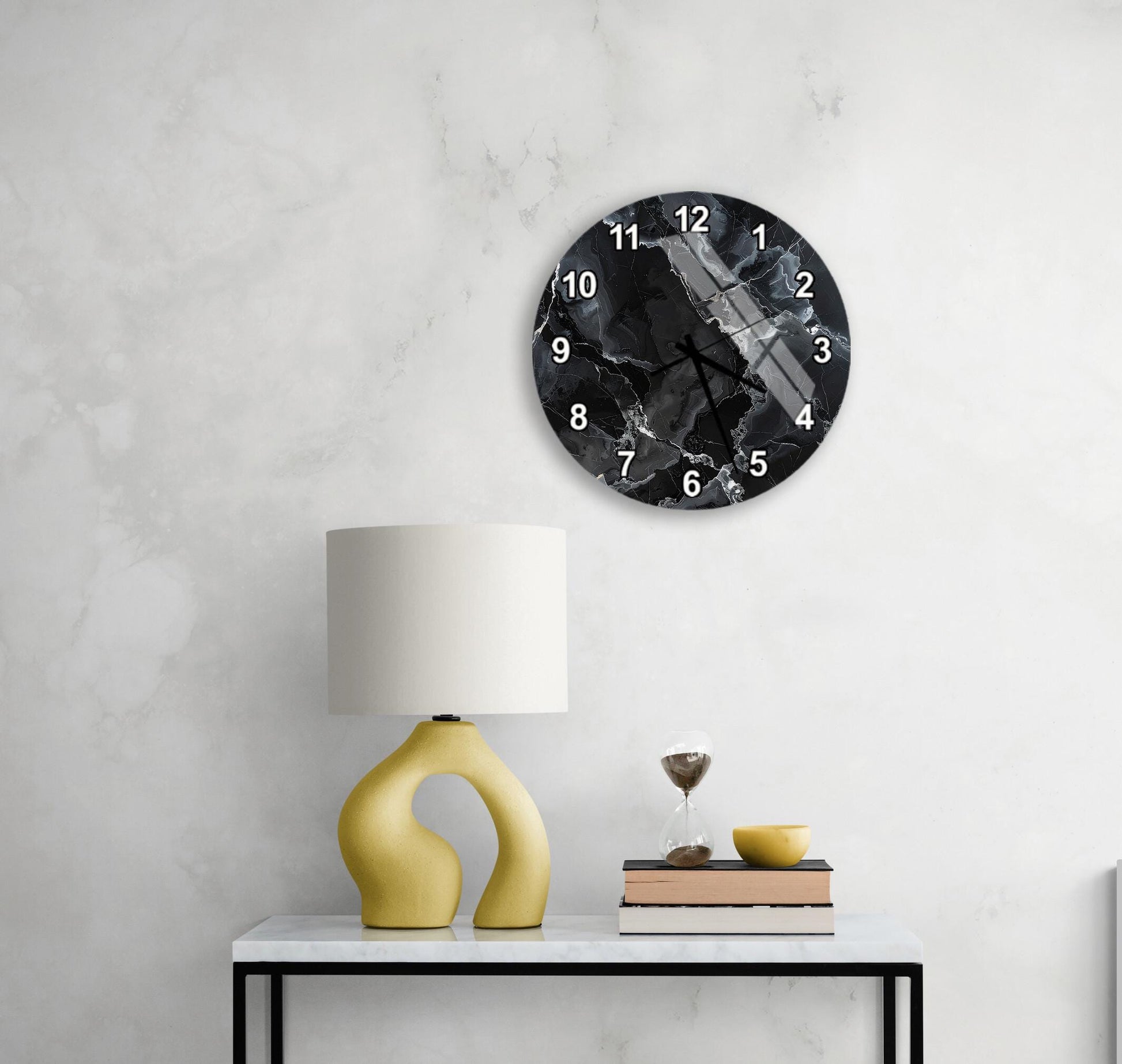 a modern black and white clock with a marble-like design, mounted on a white wall above a white table with a yellow lamp and a stack of books.