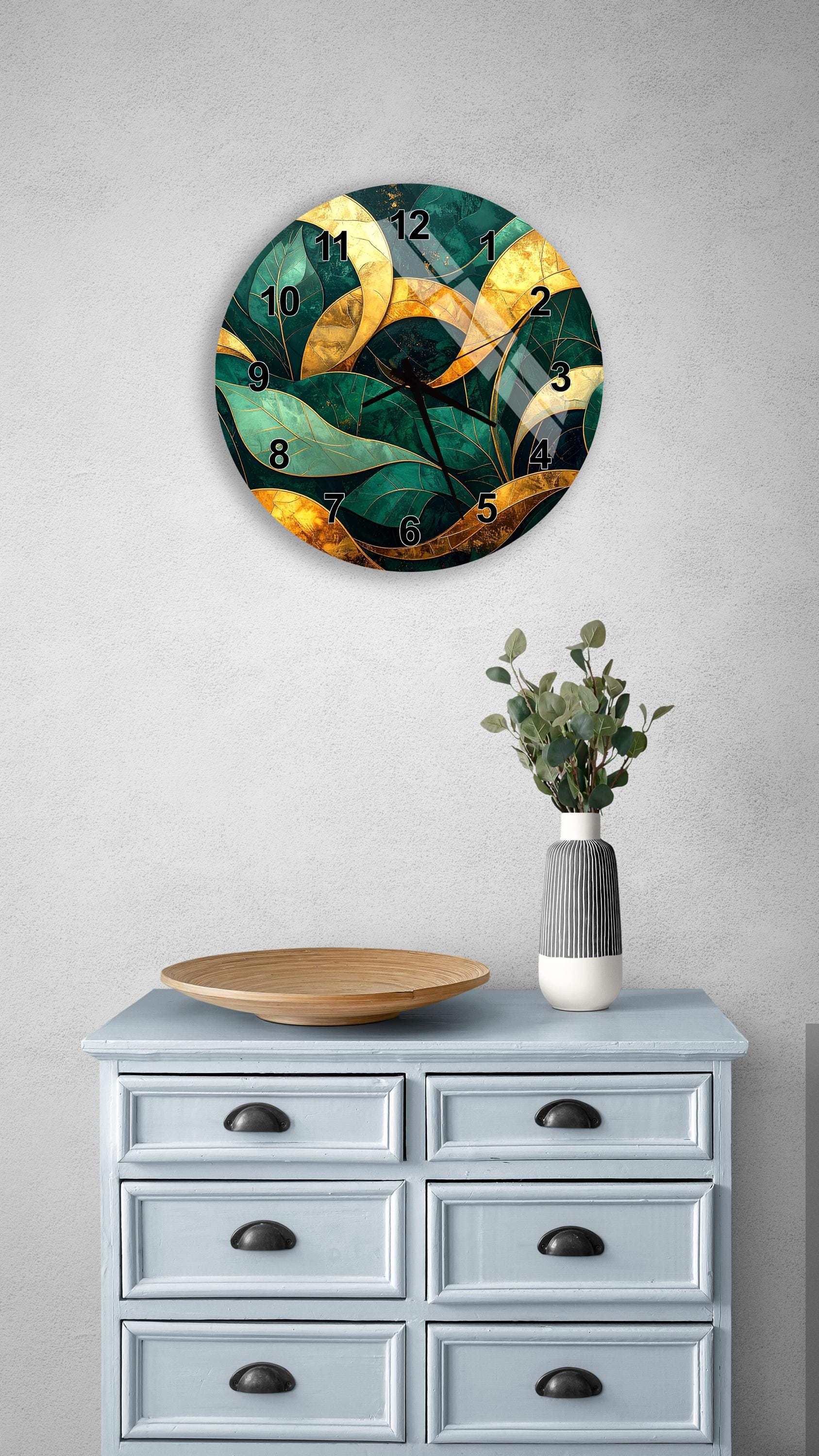 a wall clock with a green and gold leafy design, mounted on a wall above a light blue dresser with six drawers. The dresser also has a wooden plate and a vase with greenery on it.