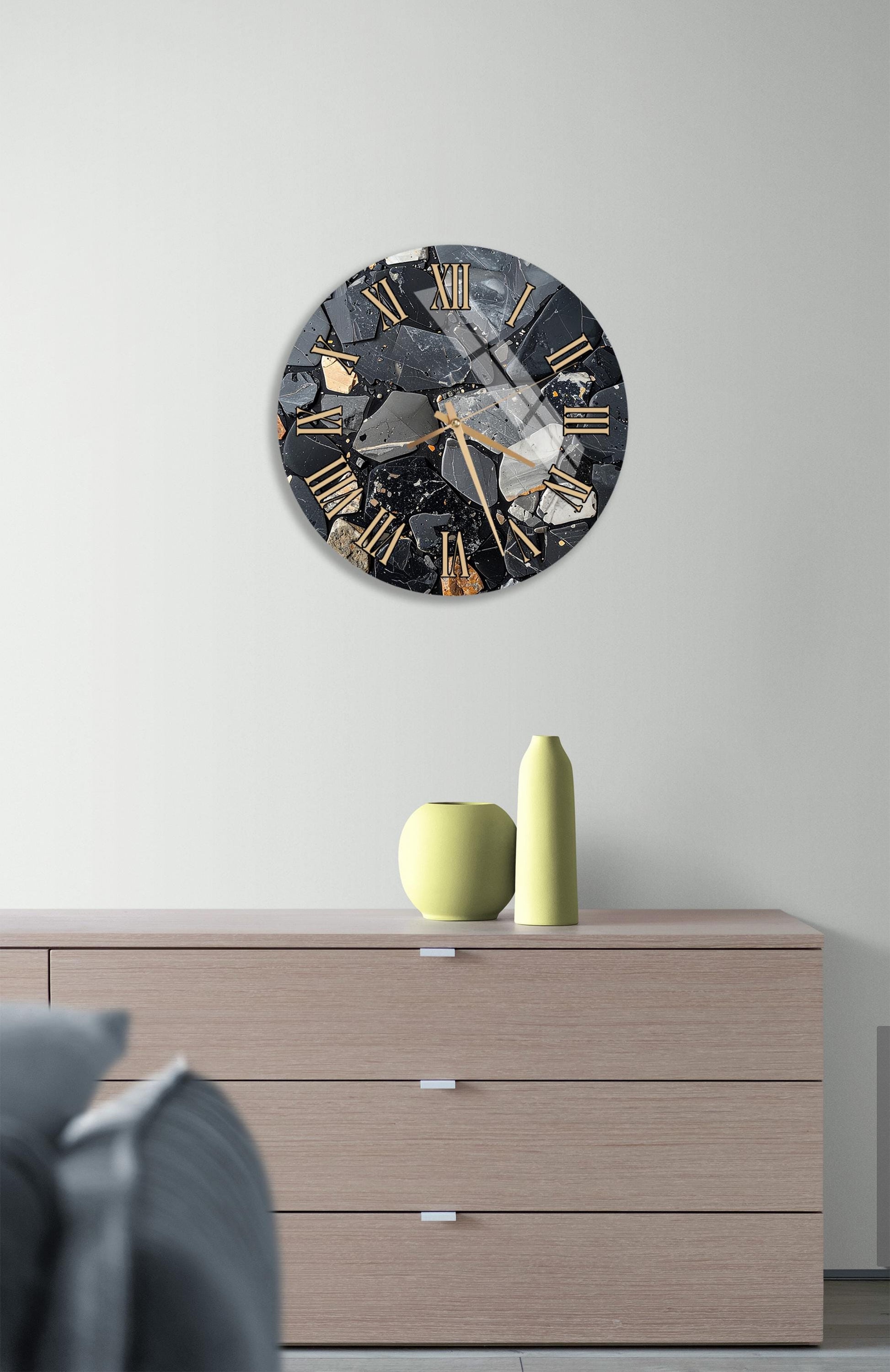 a modern clock with a black and gold design, mounted on a wall above a wooden dresser. The dresser has a vase and a small sculpture on top of it.