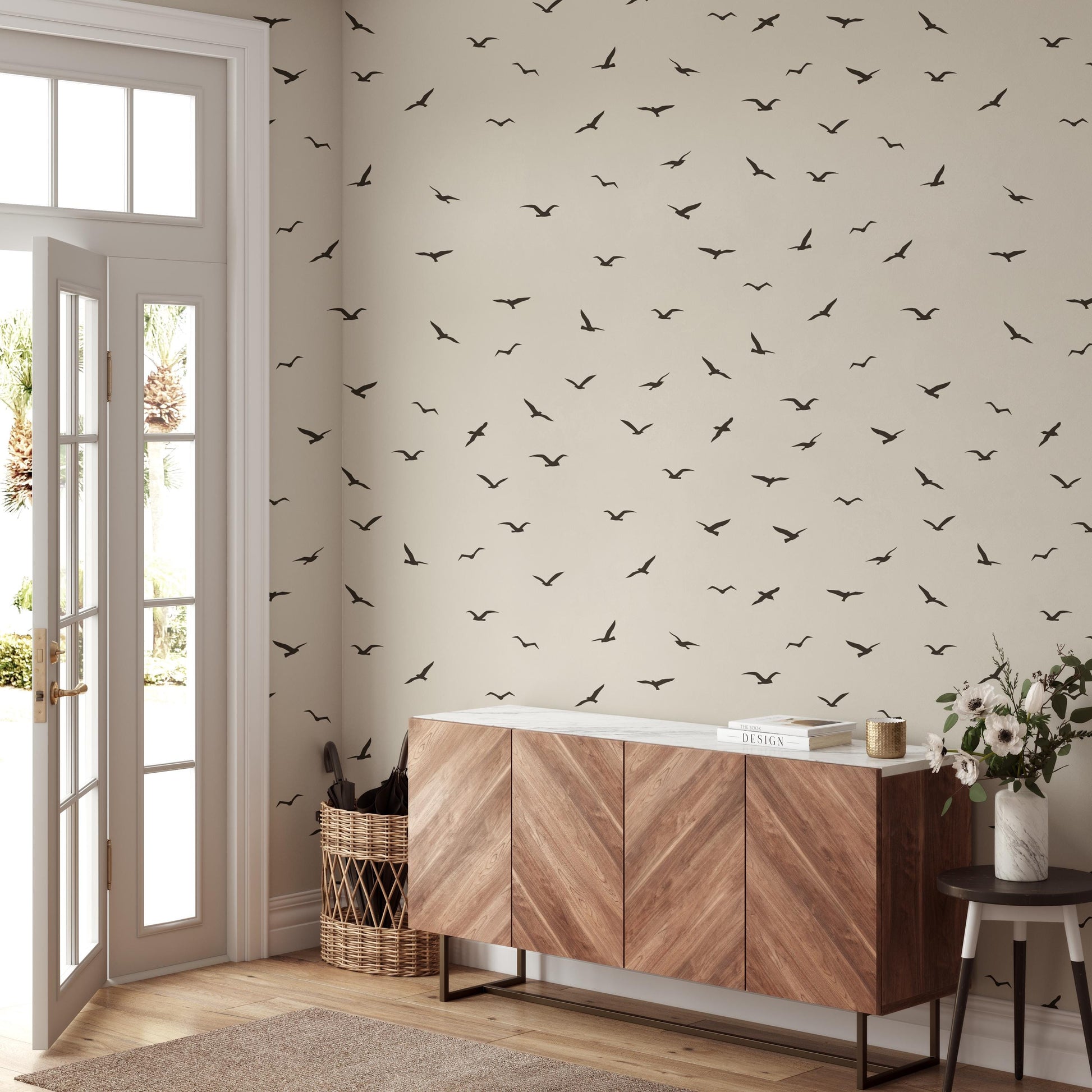 a room with a large wall covered in a pattern of black birds, creating a striking visual effect. The room is furnished with a wooden cabinet, a wicker basket, and a small table with a vase of flowers. The overall design and arrangement of the objects in the room suggest a well-organized and aesthetically pleasing space.