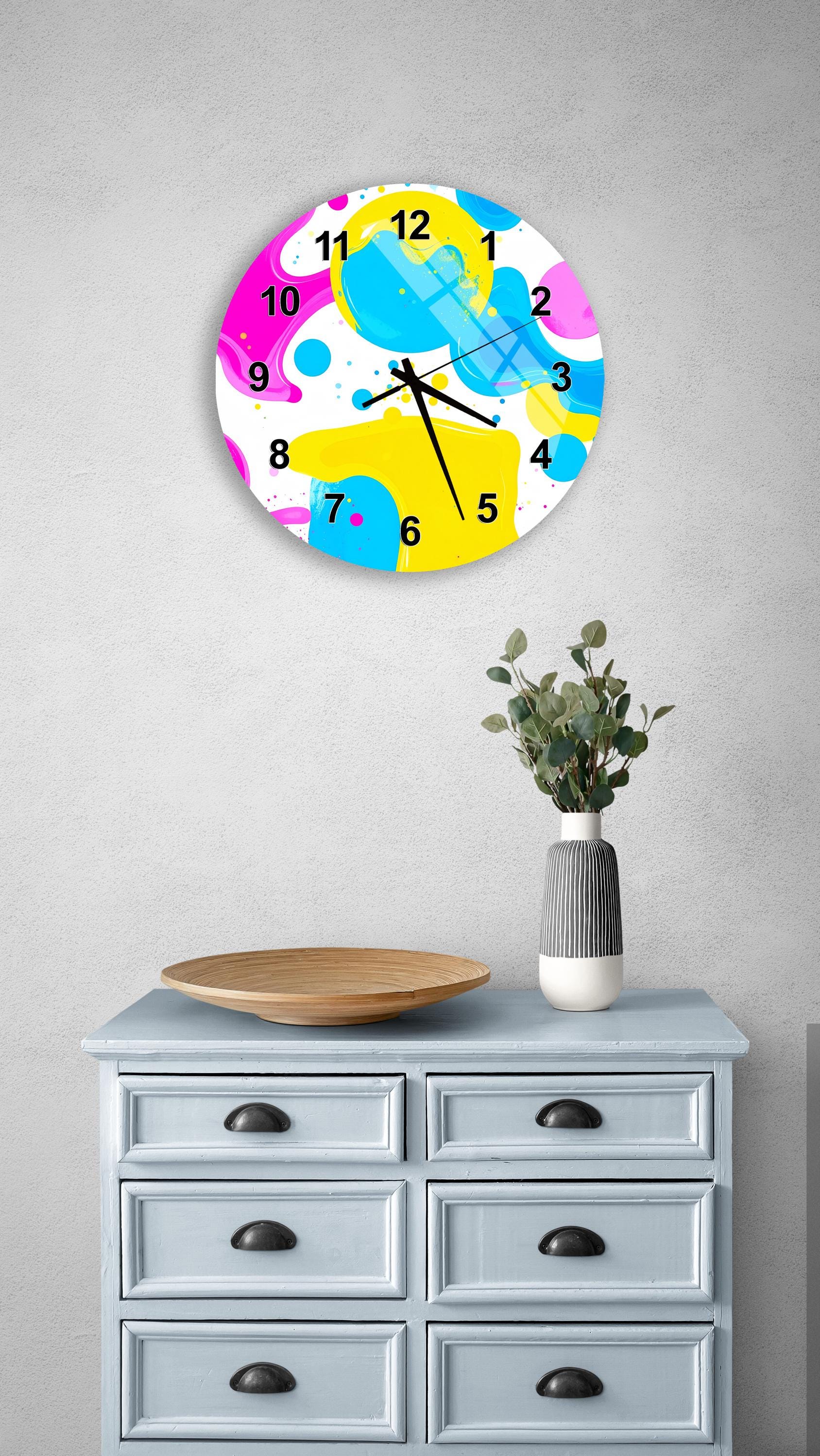 a colorful abstract clock with a vibrant design, mounted on a wall above a light blue dresser with six drawers. The dresser also has a vase of greenery and a wooden tray on top.