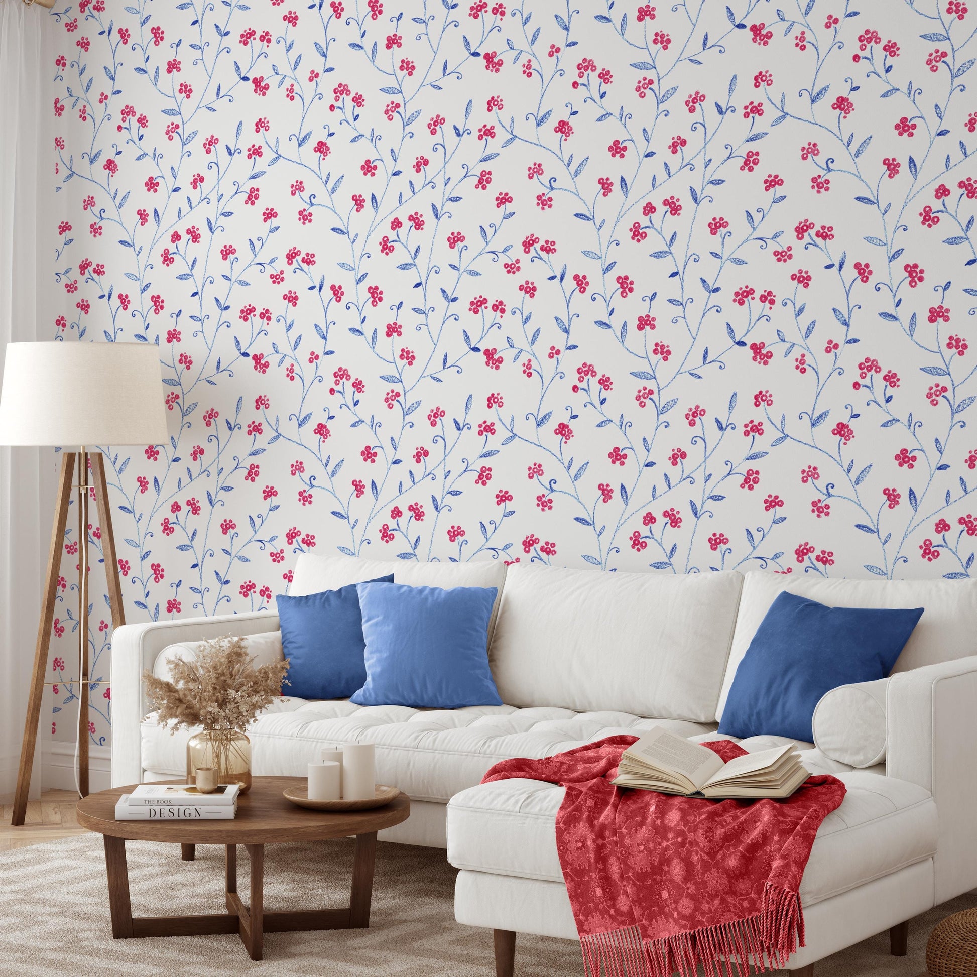 a cozy living room with a white sofa adorned with blue throw pillows, a wooden coffee table, and a lamp. The walls are decorated with a floral wallpaper, and a red blanket is draped over the sofa.