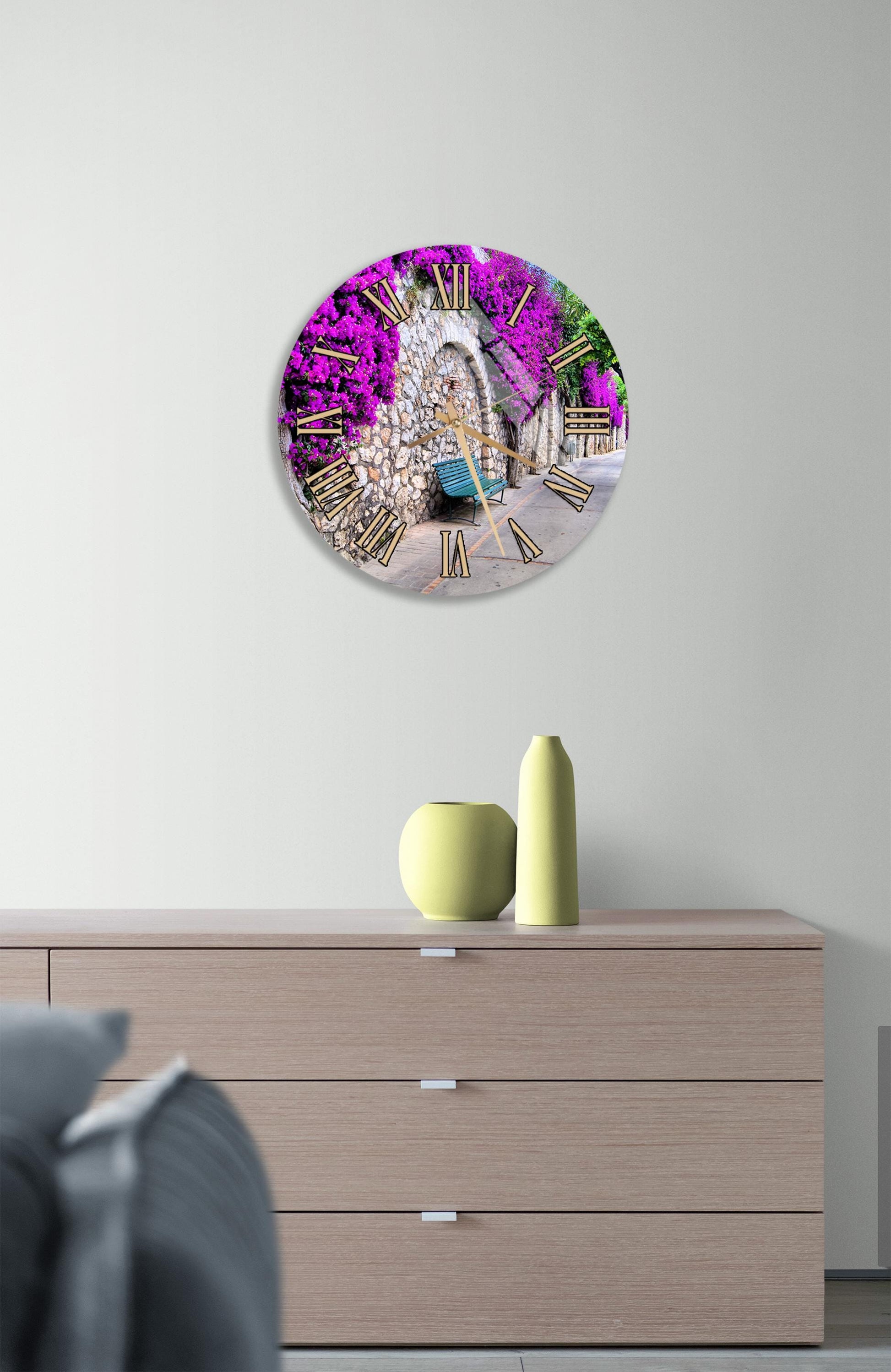 a round clock with a vibrant purple and pink floral design, mounted on a wall above a wooden dresser with a vase and a small sculpture on top.
