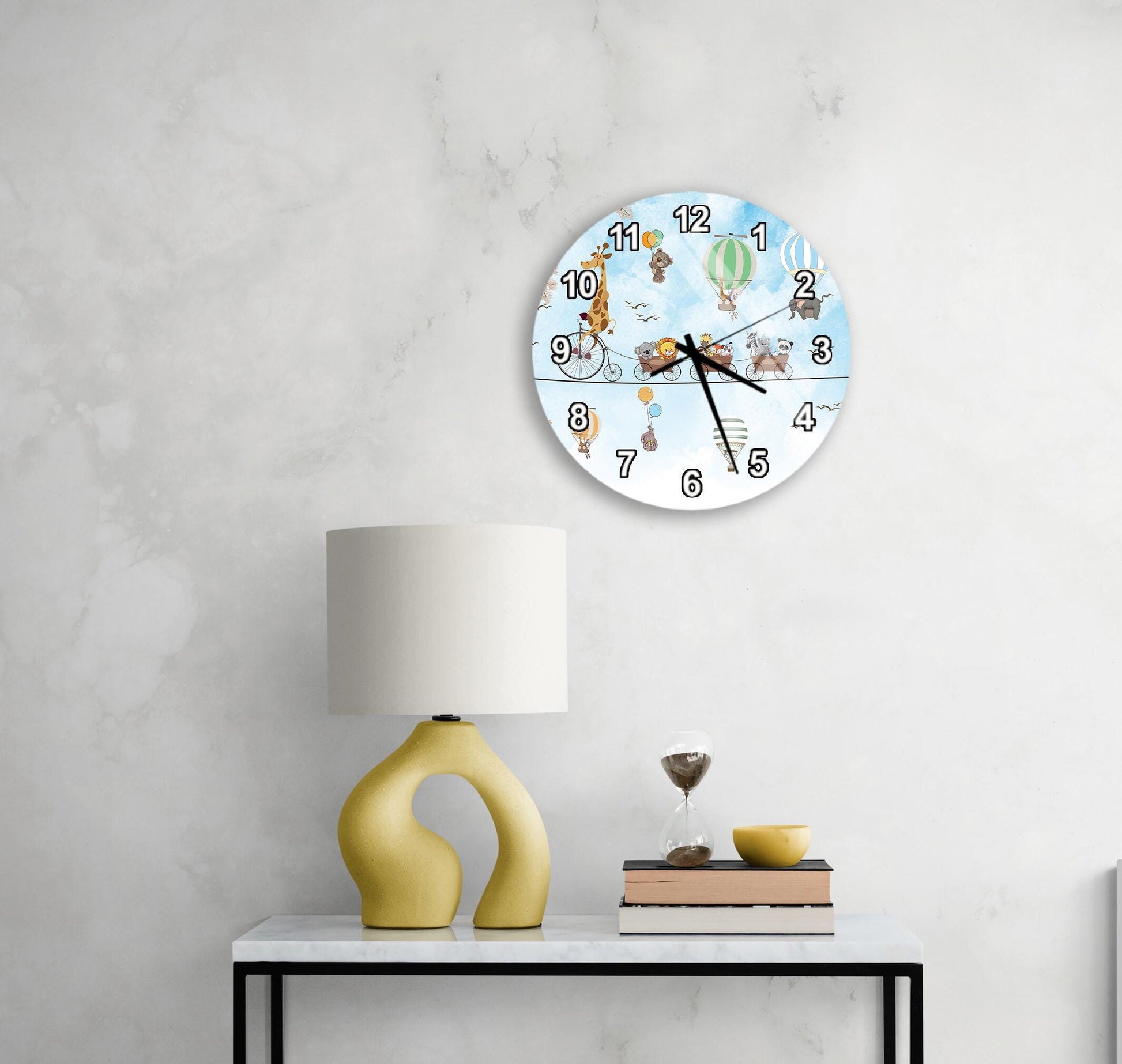 a round clock with a cartoon-style illustration of a giraffe and a hot air balloon on its face, mounted on a wall above a white table with a yellow lamp and a stack of books.