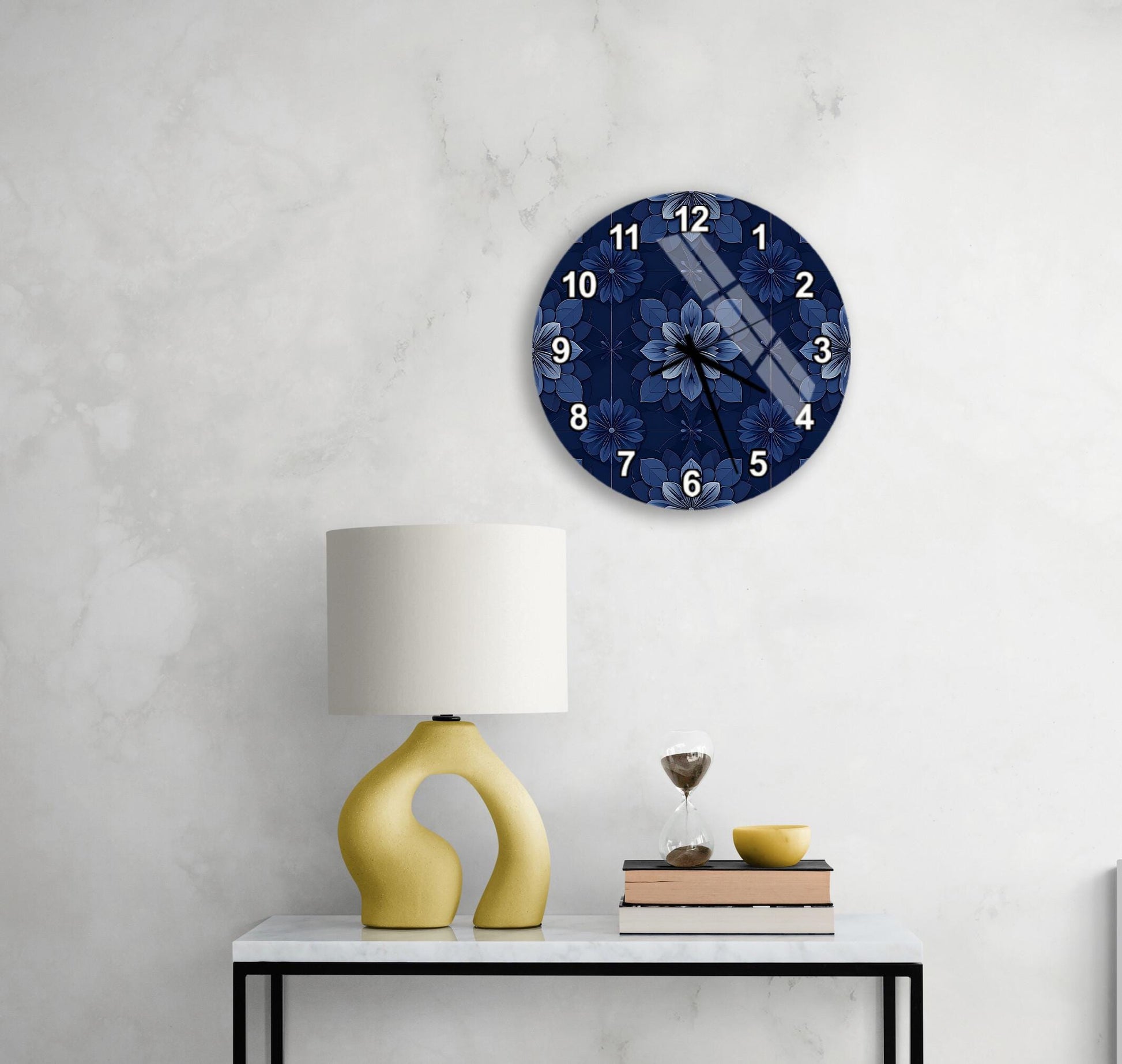 a white marble wall with a round clock displaying a blue floral pattern, a yellow lamp with a unique curved base, a small table with a book and a glass of water, and a stack of books.