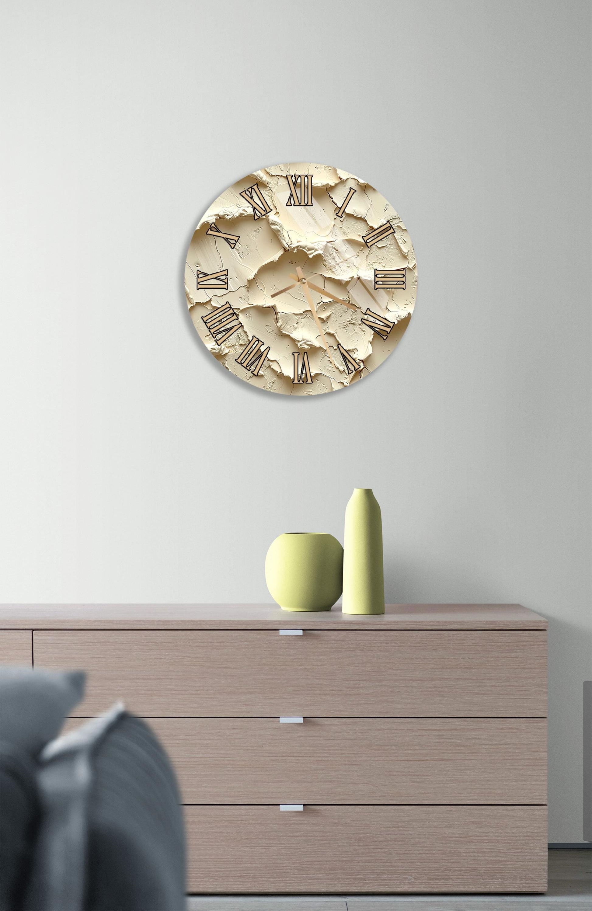 a wall clock with a distressed beige finish, mounted on a wall above a wooden dresser with three drawers. The dresser also has a small green vase and a yellow vase placed on it.