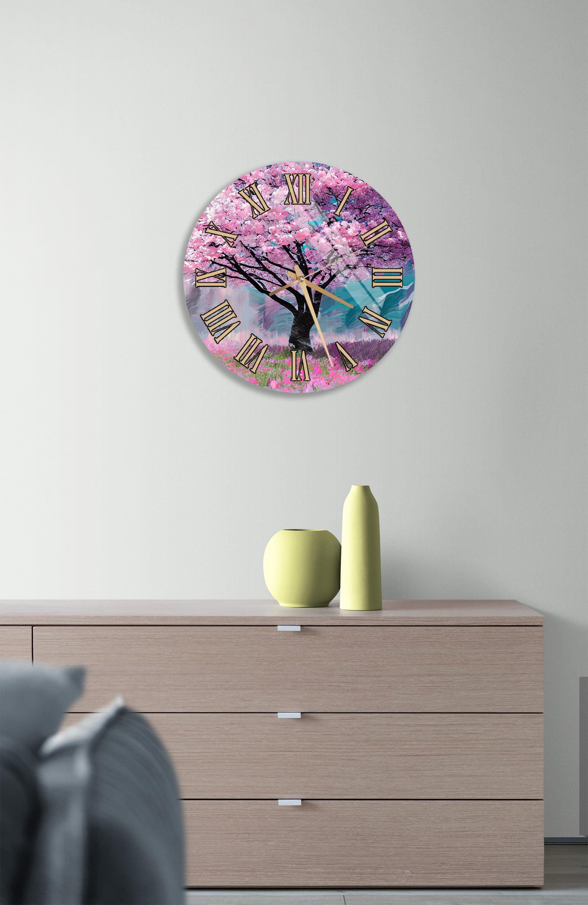 a wall clock with a pink tree design, mounted on a wall above a wooden dresser with a vase and a small sculpture on top.