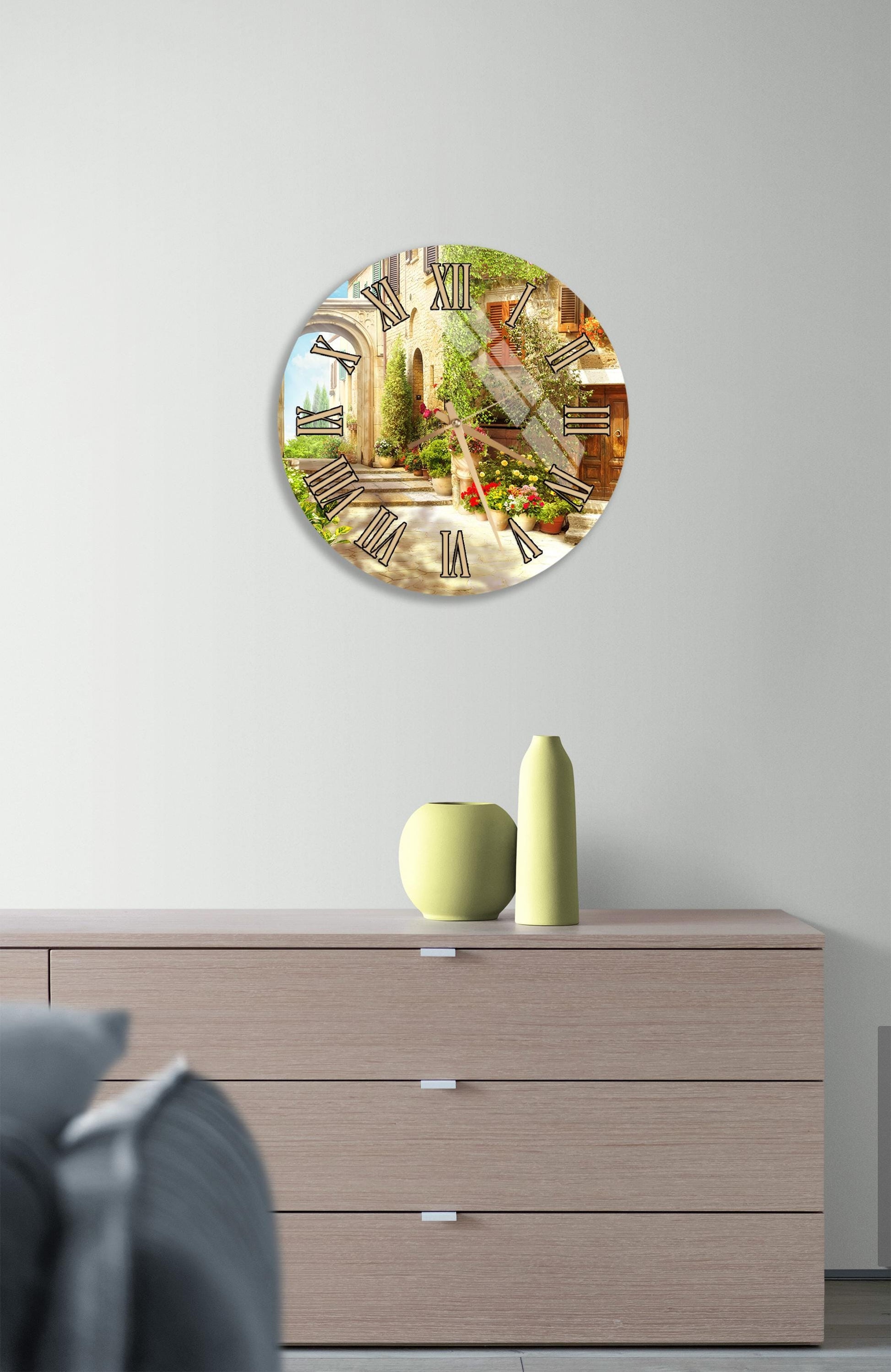 a wall clock with a scenic background, a wooden dresser with a vase and a small sculpture, and a gray couch in the foreground.