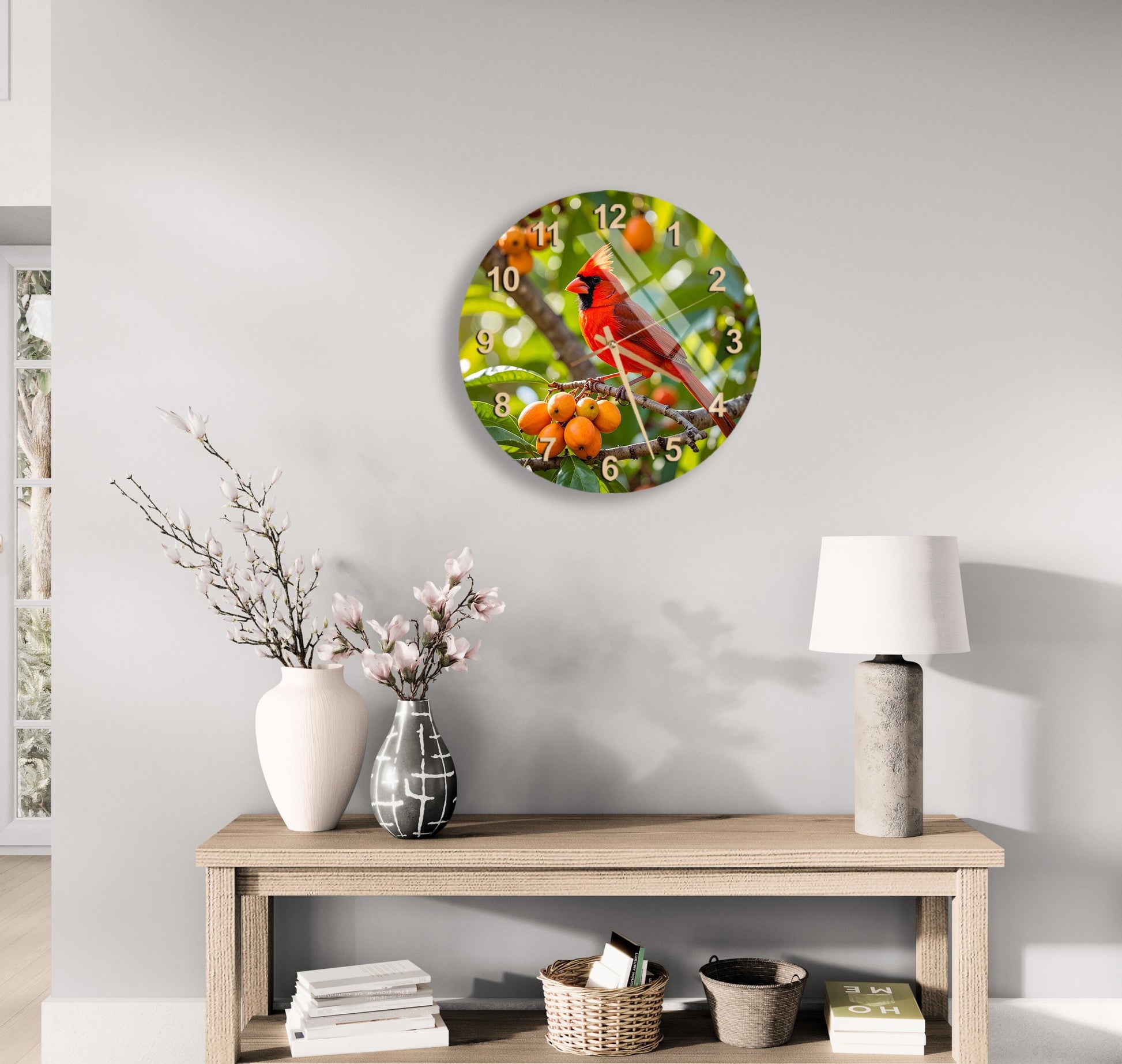 a wall clock with a red cardinal bird image on it, hanging above a wooden table with a lamp, a vase with flowers, and some books.