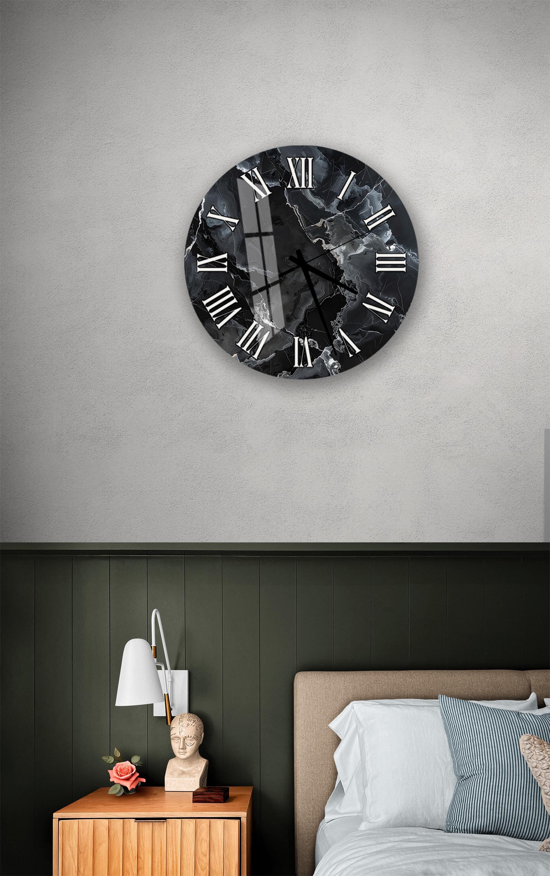 a bedroom setting with a large black and white clock on the wall above a bed. The clock has a marble-like design and Roman numerals. The room has a green wall, a wooden nightstand with a lamp, and a white headboard.