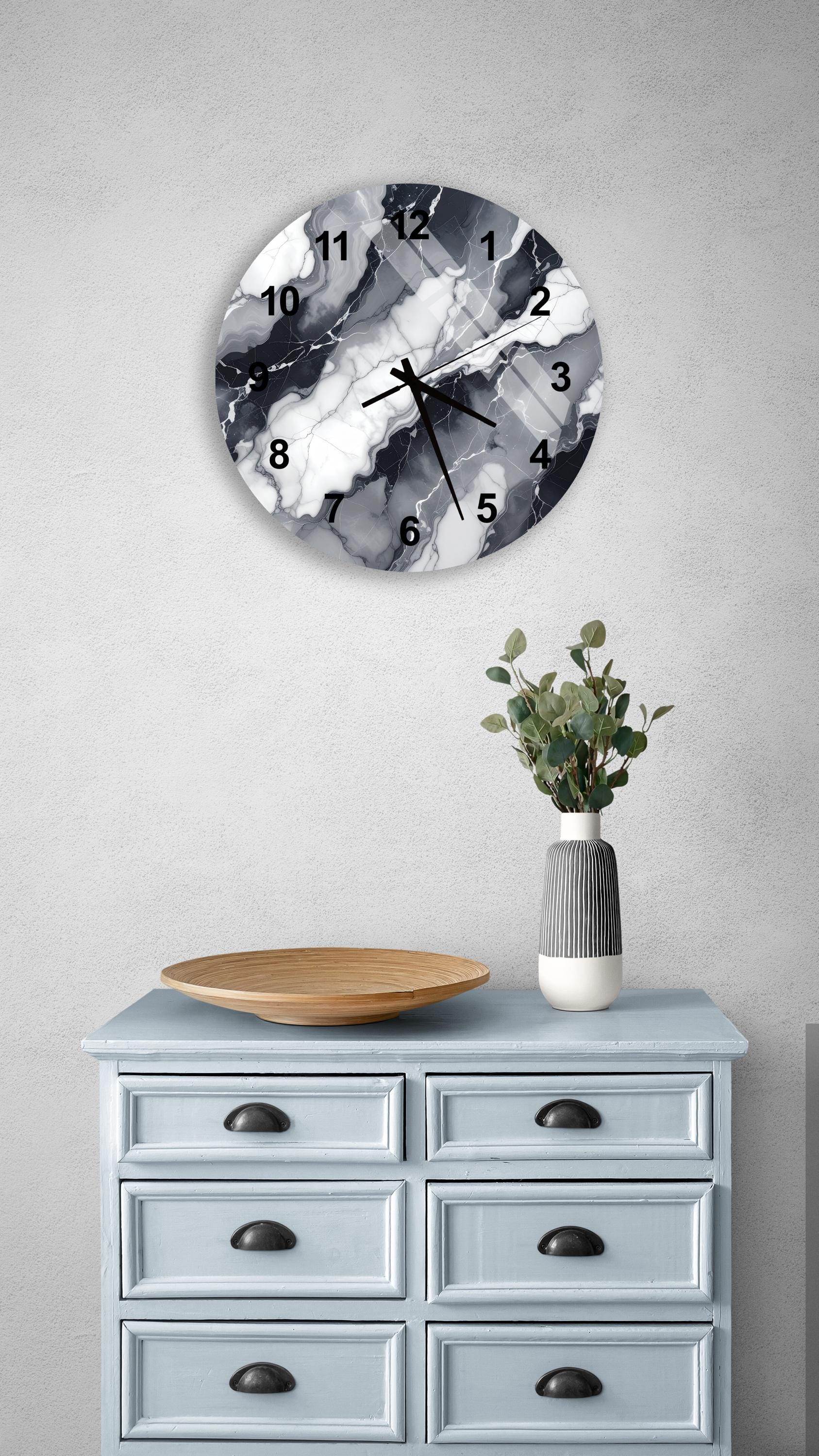 a wall clock with a marble-like design, mounted on a wall above a light blue dresser with six drawers. The dresser also has a vase with greenery on it, and a wooden bowl on top.