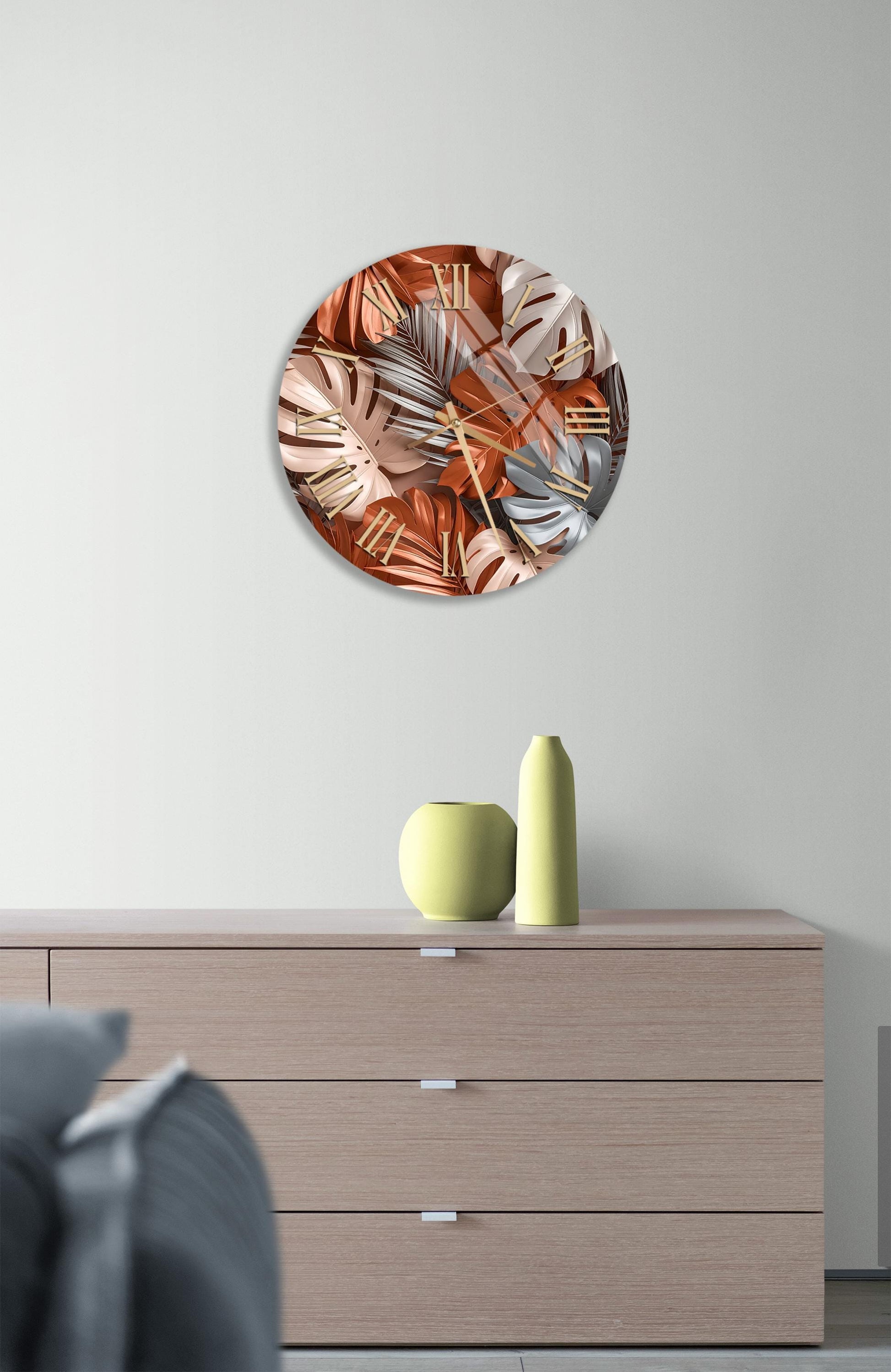 a round wall art piece with a vibrant design of orange and pink leaves, hanging above a wooden dresser with a yellow vase and a gray vase on top.