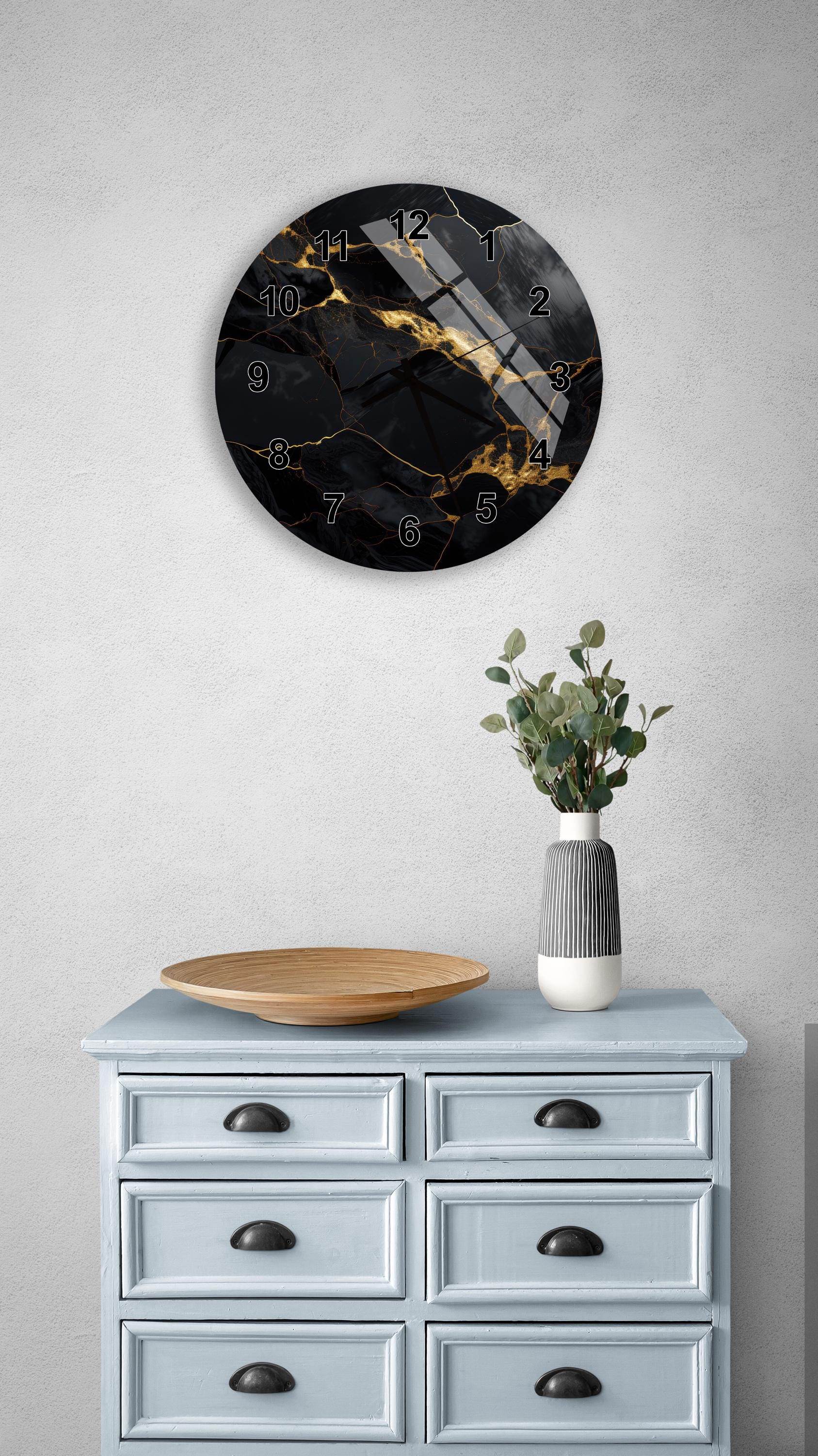 a wall clock with a black and gold design, mounted on a wall above a light blue dresser with six drawers. The dresser also has a vase with greenery and a wooden bowl on it.
