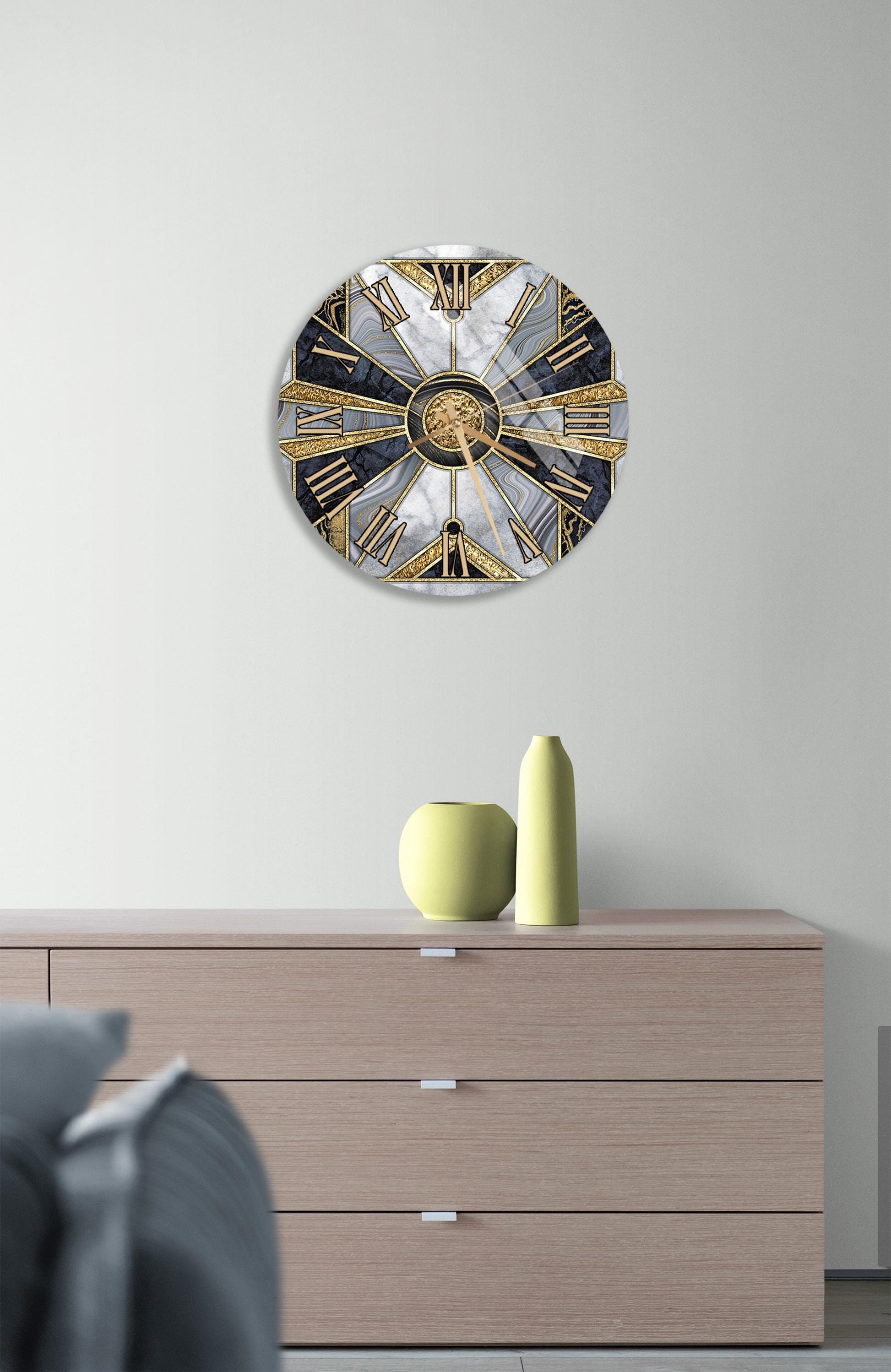 a modern interior design with a large, round clock with a gold and black design on a white wall above a wooden dresser. The dresser has a vase and a small sculpture on top of it.