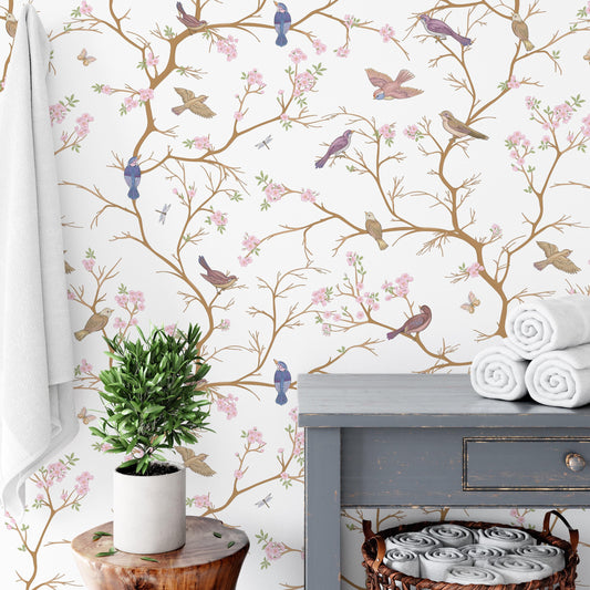 a bathroom setting with a white towel, a potted plant, and a wooden table with rolled-up towels on it. The wall behind the objects is decorated with a floral wallpaper pattern of birds and flowers.