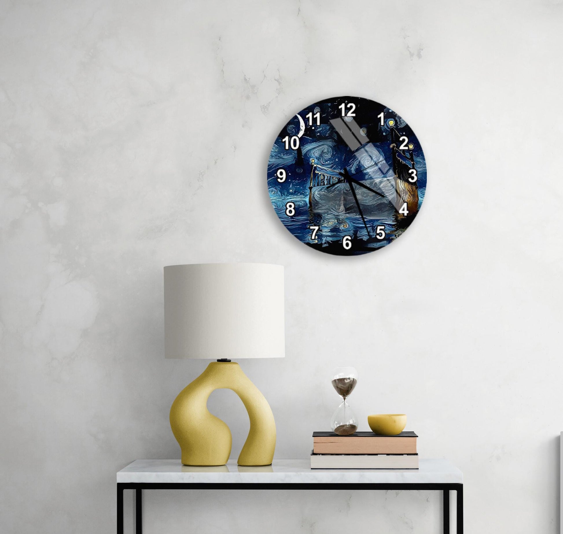 a modern wall clock with a blue and white design, mounted on a white wall above a white table with a yellow lamp and a stack of books.