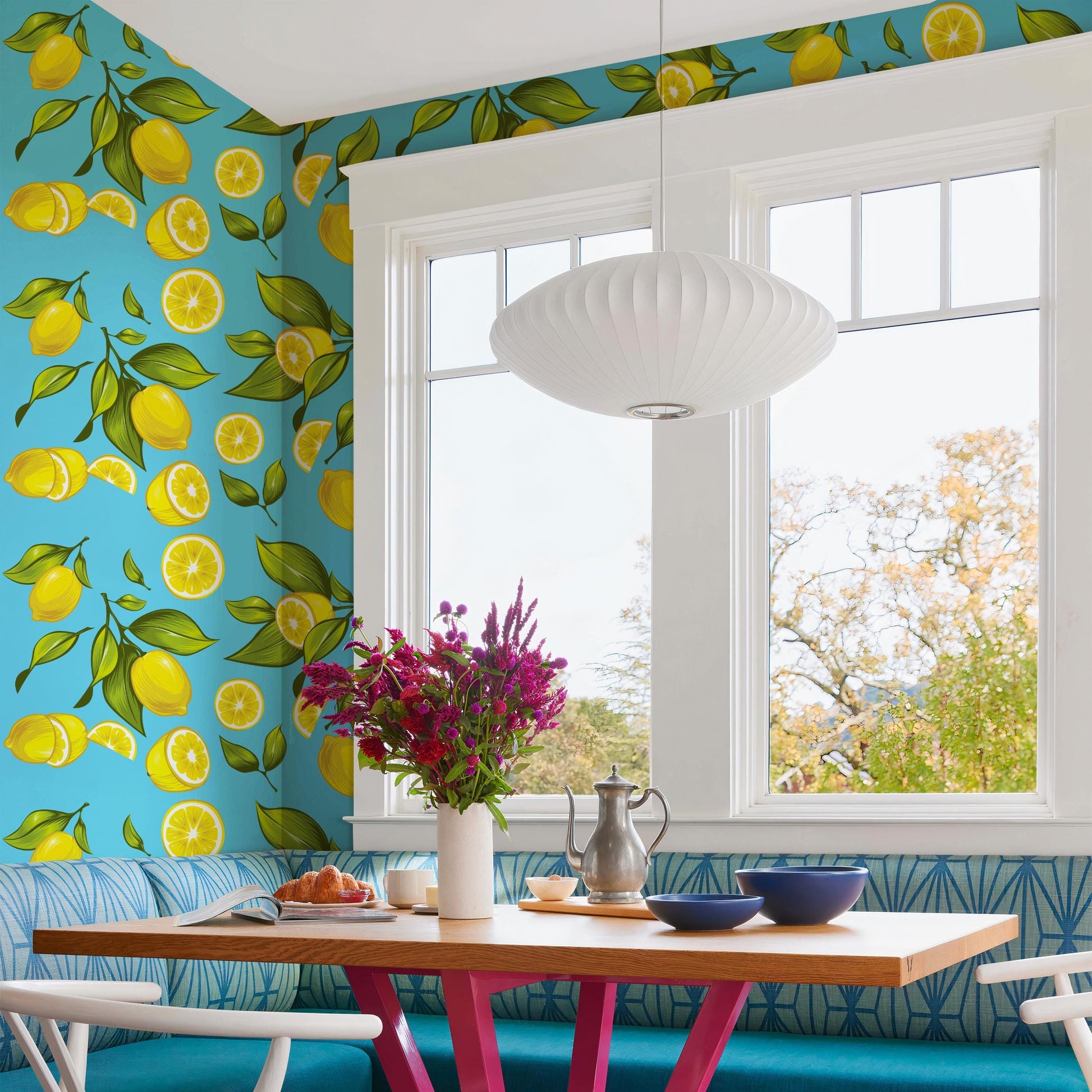 a bright and colorful kitchen with a large window overlooking a lush green outdoor scene. The kitchen has a blue and white color scheme, with a wooden table and chairs, a vase of purple flowers, and a pendant light hanging above the table.