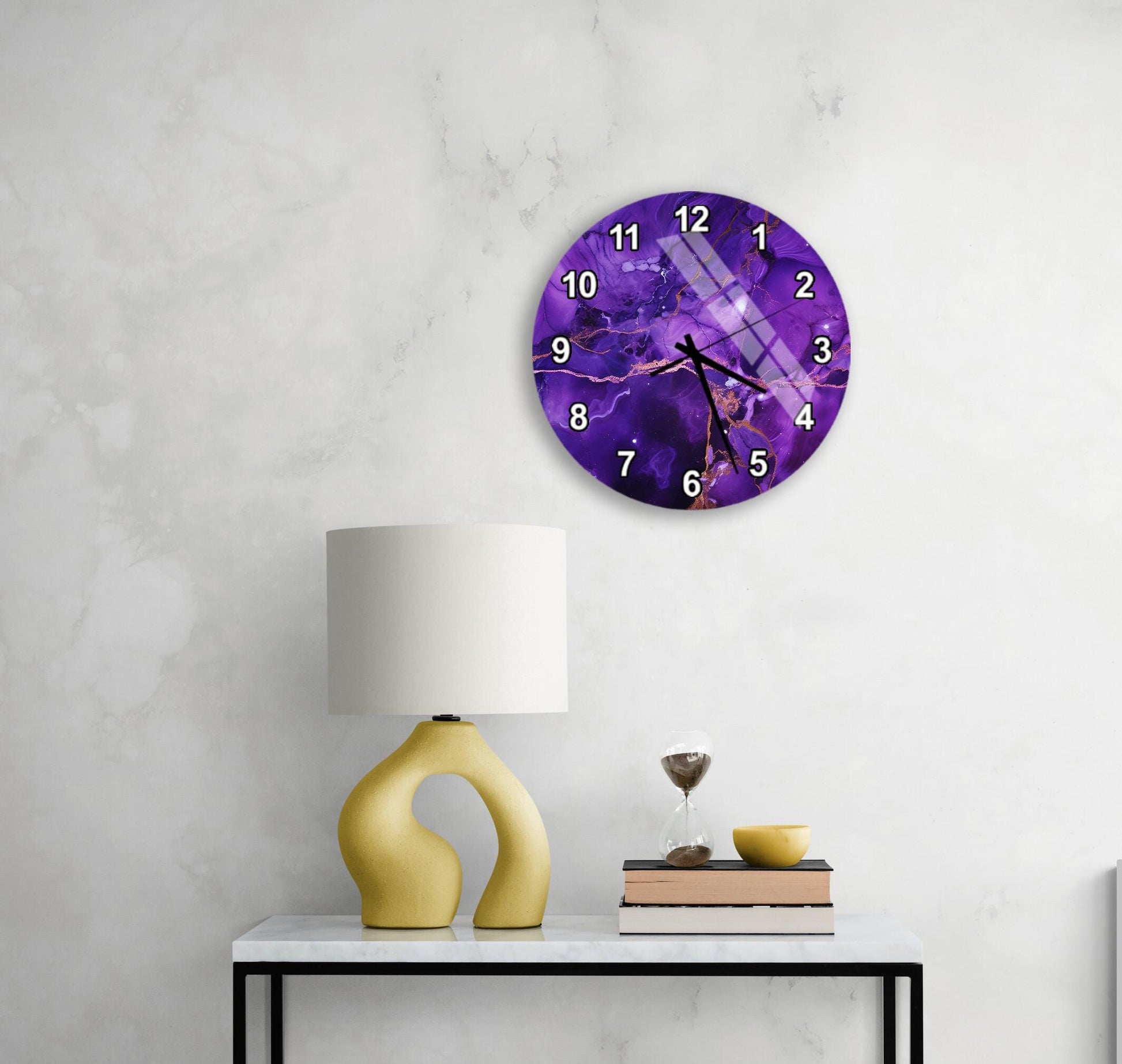 a white wall with a purple clock featuring a purple and white design, a yellow lamp, a stack of books, and a glass of wine on a white table.