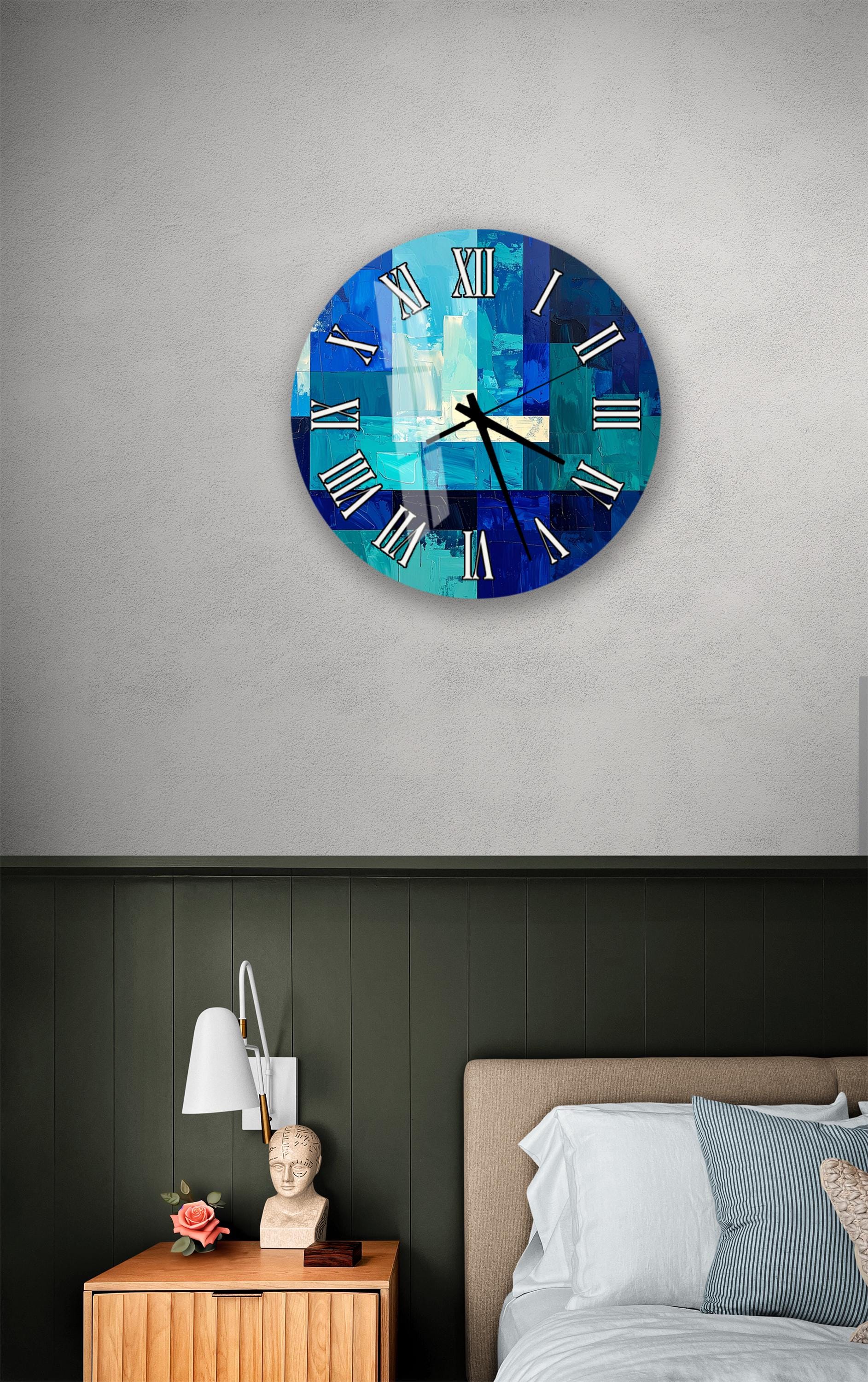 a bedroom setting with a large blue and white clock on the wall above a bed. The clock has a unique abstract design and is mounted on a green wall. The bed has a white headboard and a wooden nightstand with a lamp and a vase of flowers.