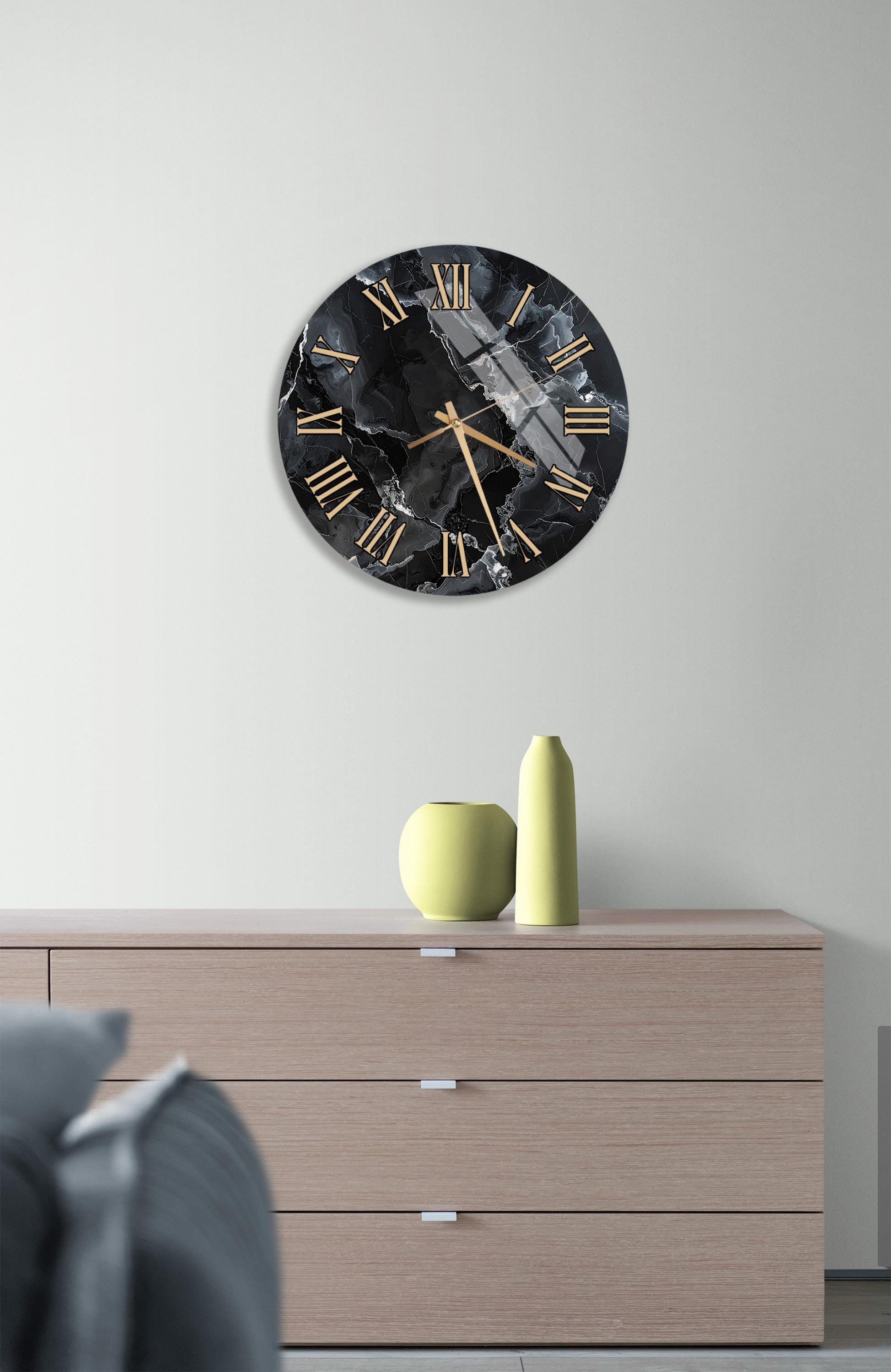 a modern black and gold clock with Roman numerals on a white wall, mounted above a wooden dresser with a yellow vase and a gray couch in the foreground.