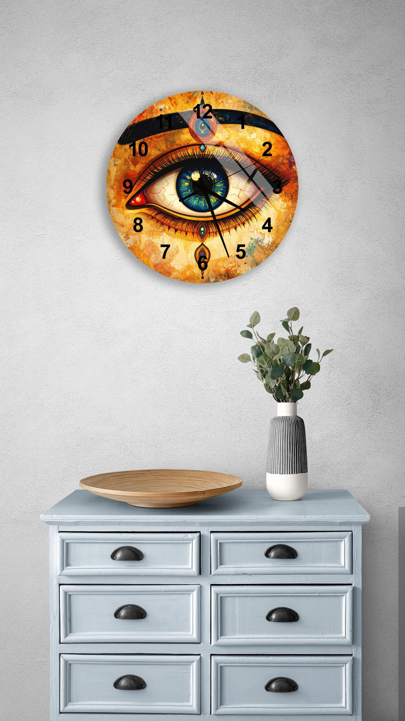 a wall clock with an eye design, mounted on a wall above a wooden dresser with drawers. The dresser also has a vase with greenery and a wooden tray on it.
