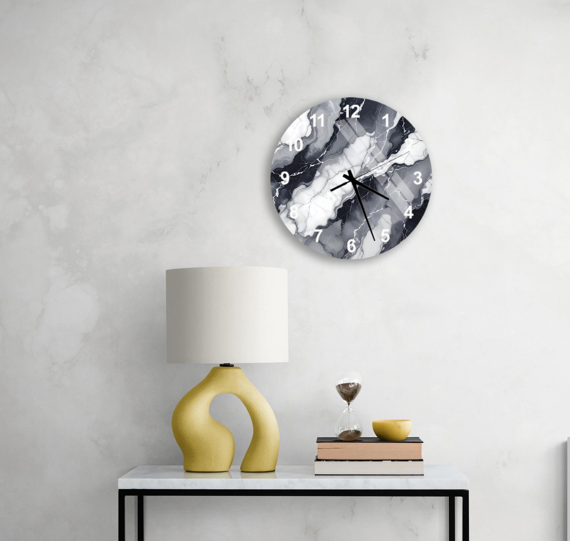 a modern black and white clock with a marble-like design, mounted on a wall above a white table with a yellow lamp and a stack of books.