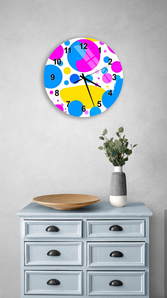 a colorful clock with a polka dot design, mounted on a wall above a wooden dresser with six drawers. The dresser also has a vase with greenery on it, and there is a wooden plate on top of it.