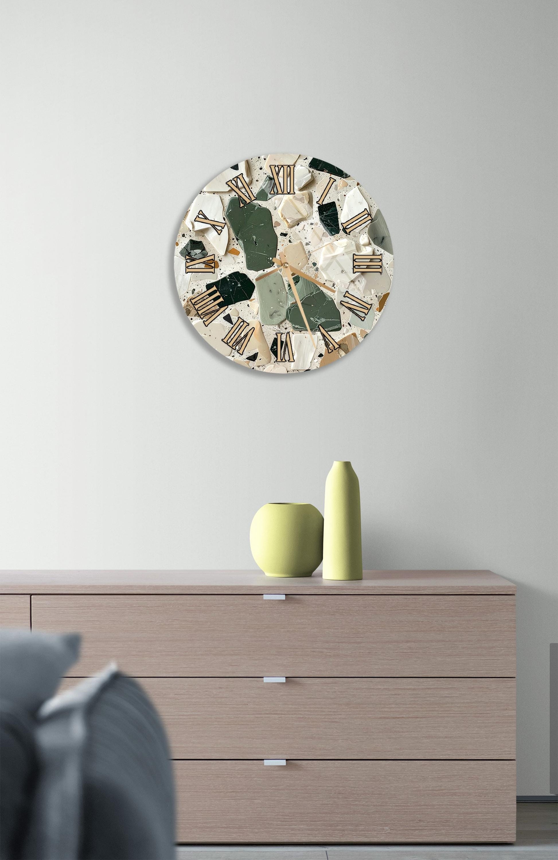 a modern interior design with a round clock featuring a mosaic pattern on the wall above a wooden dresser with three drawers. The dresser also has two yellow vases and a gray couch in the background.