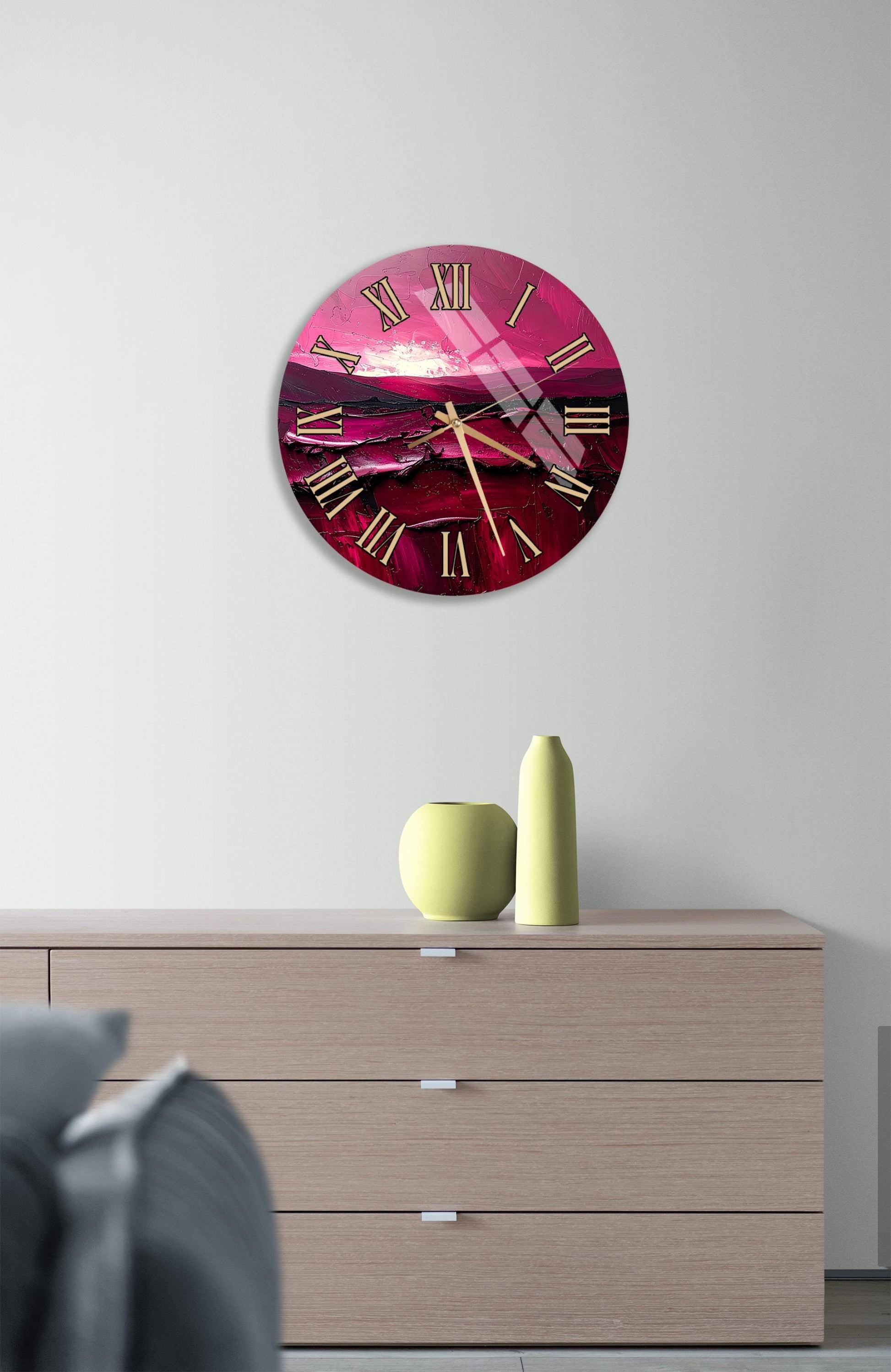 a large, round clock with a pink and purple color scheme, mounted on a wall above a wooden dresser. The dresser has a few decorative items on it, including a yellow vase and a green vase.
