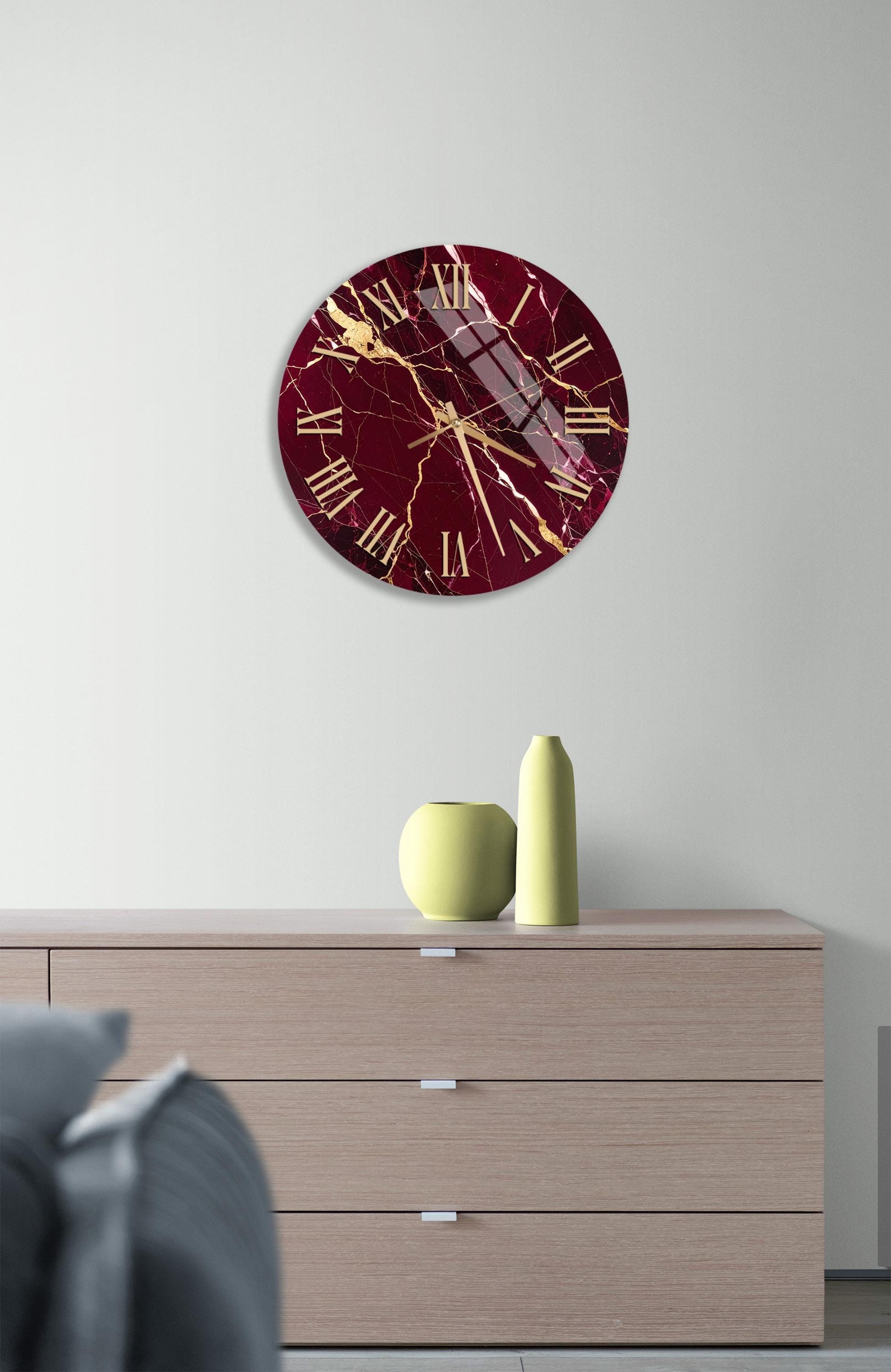 a modern interior design with a large burgundy-colored clock with gold accents hanging on a white wall above a wooden dresser. The dresser has three drawers and a small yellow vase on top.