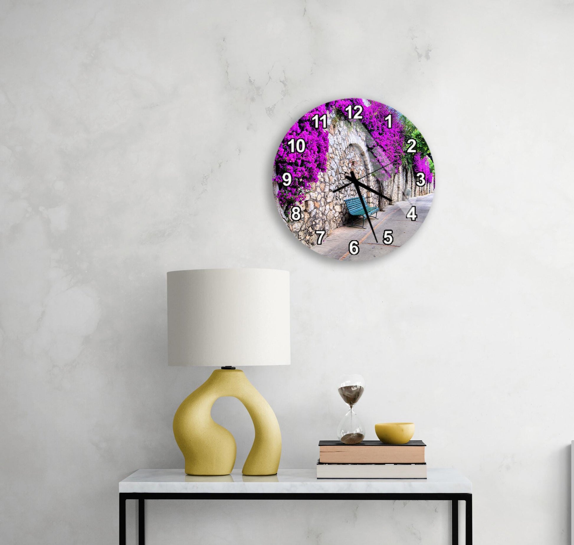 a round clock with a purple floral design on the wall above a white table with a yellow lamp and a stack of books.