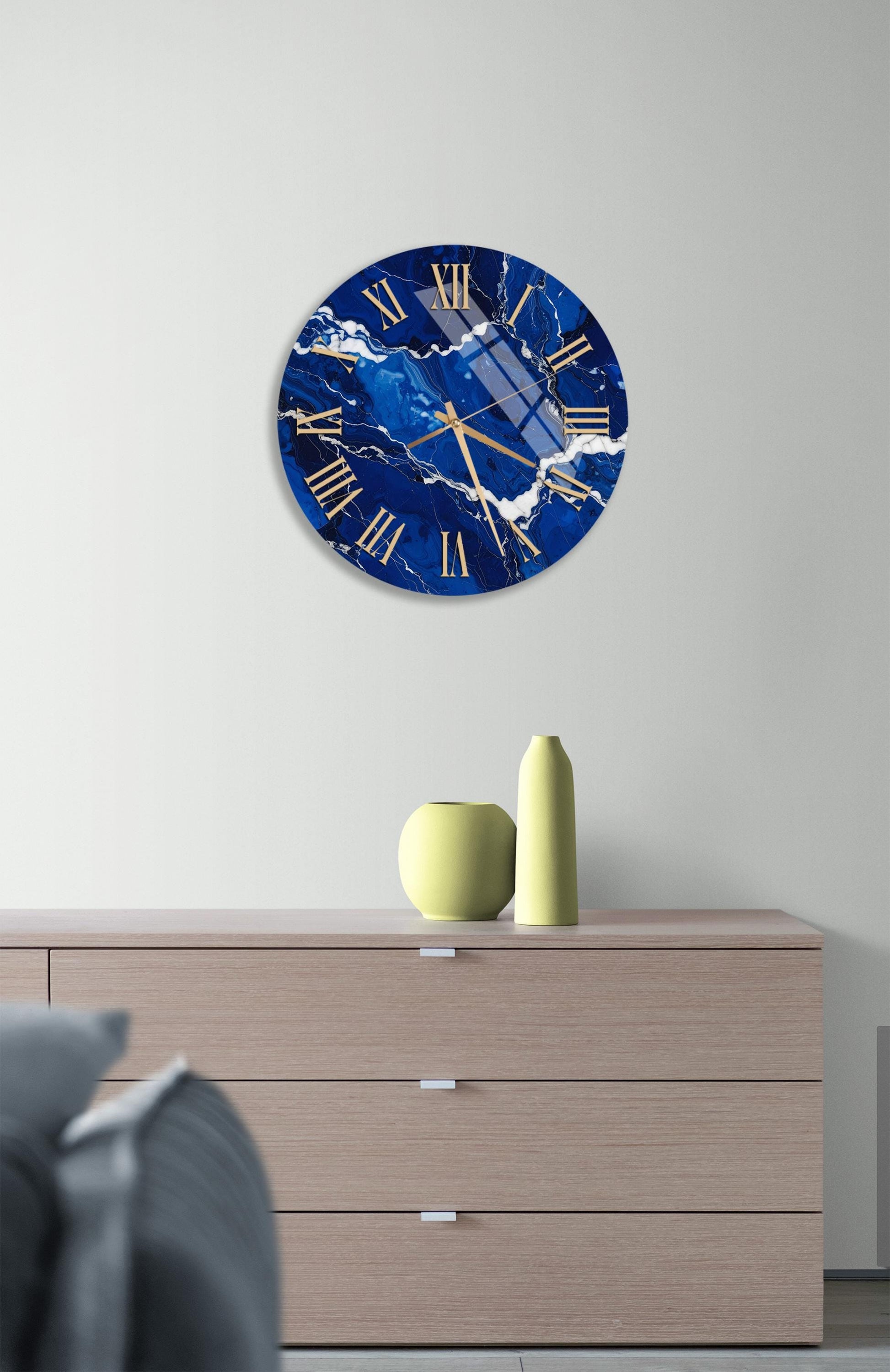a modern interior design with a large blue marble clock hanging on a white wall above a wooden dresser. The dresser has three drawers and a yellow vase on top.