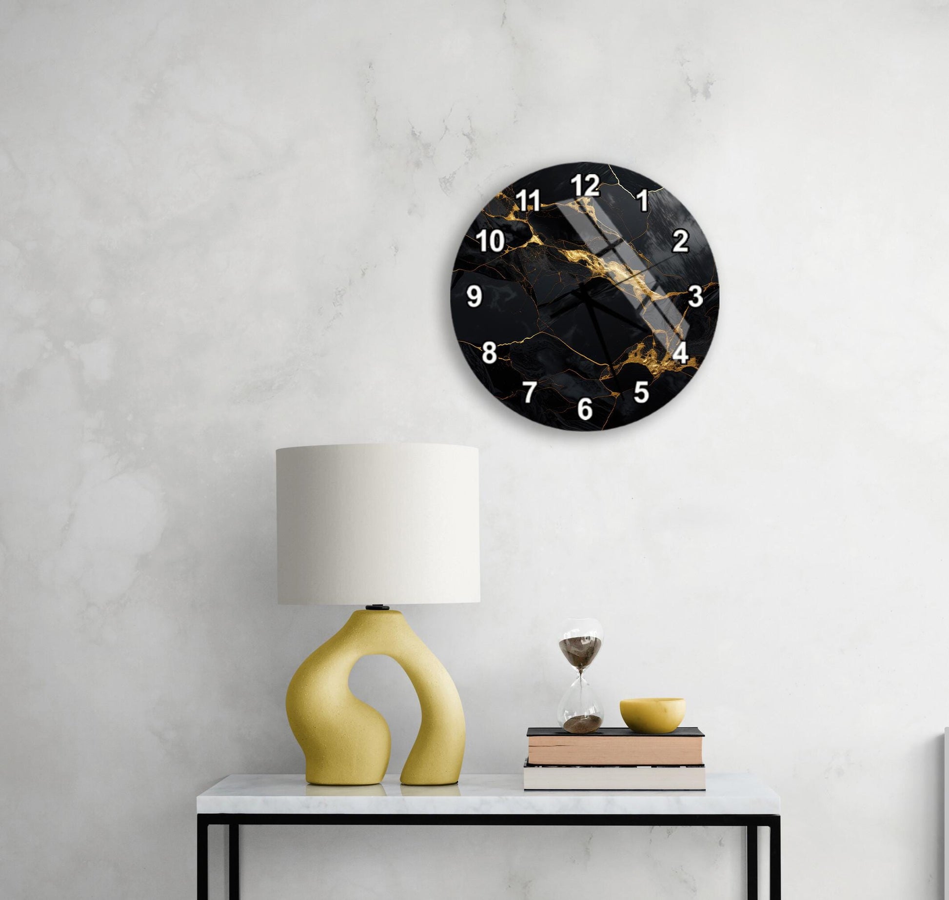 a modern black and gold wall clock with a white base, hanging on a white wall above a white table with a yellow lamp and a stack of books.