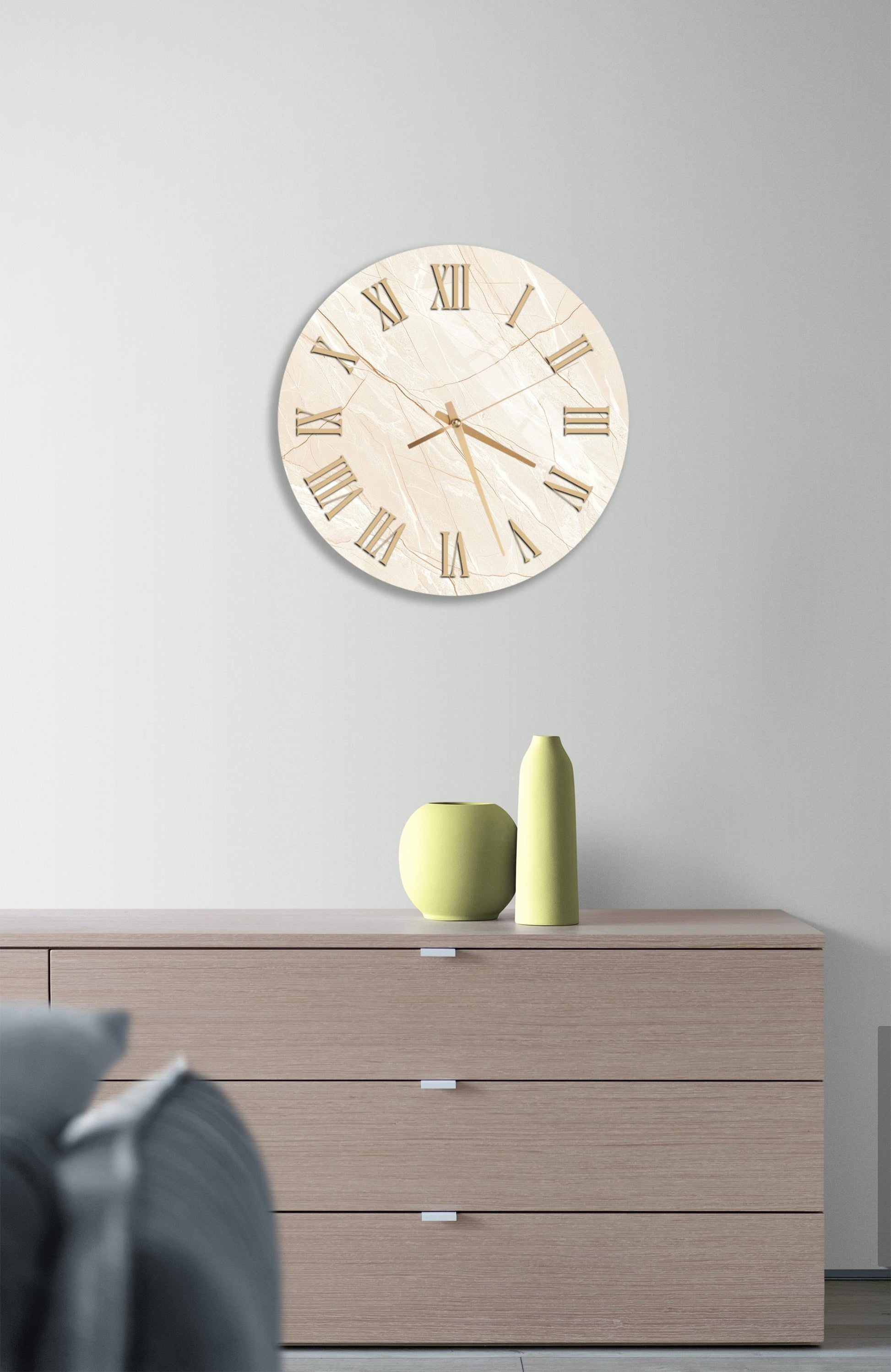 a modern living room with a large, round clock hanging on the wall above a wooden dresser. The dresser has three drawers and a vase on top. The room has a neutral color scheme, with a gray couch and a gray rug.
