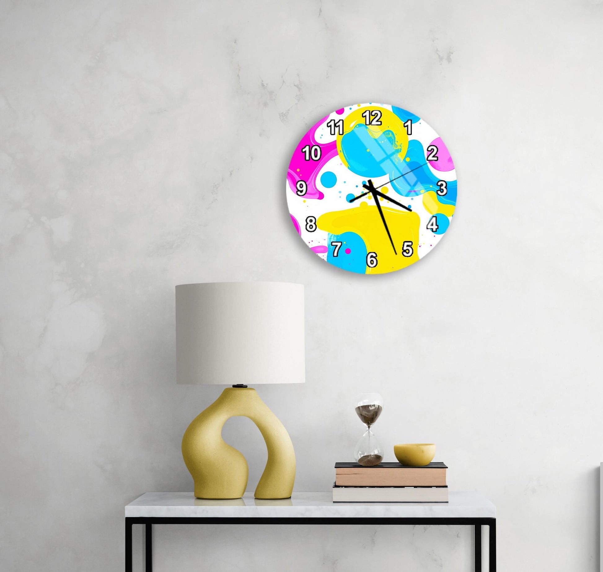 a wall clock with a colorful, abstract design hanging on a wall above a white table with a yellow lamp and a stack of books.