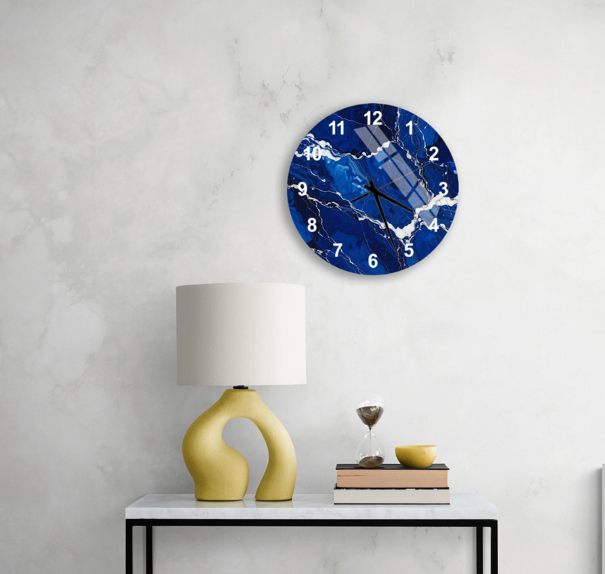 a modern wall clock with a blue marble design, mounted on a white wall above a white table with a yellow lamp and a stack of books.