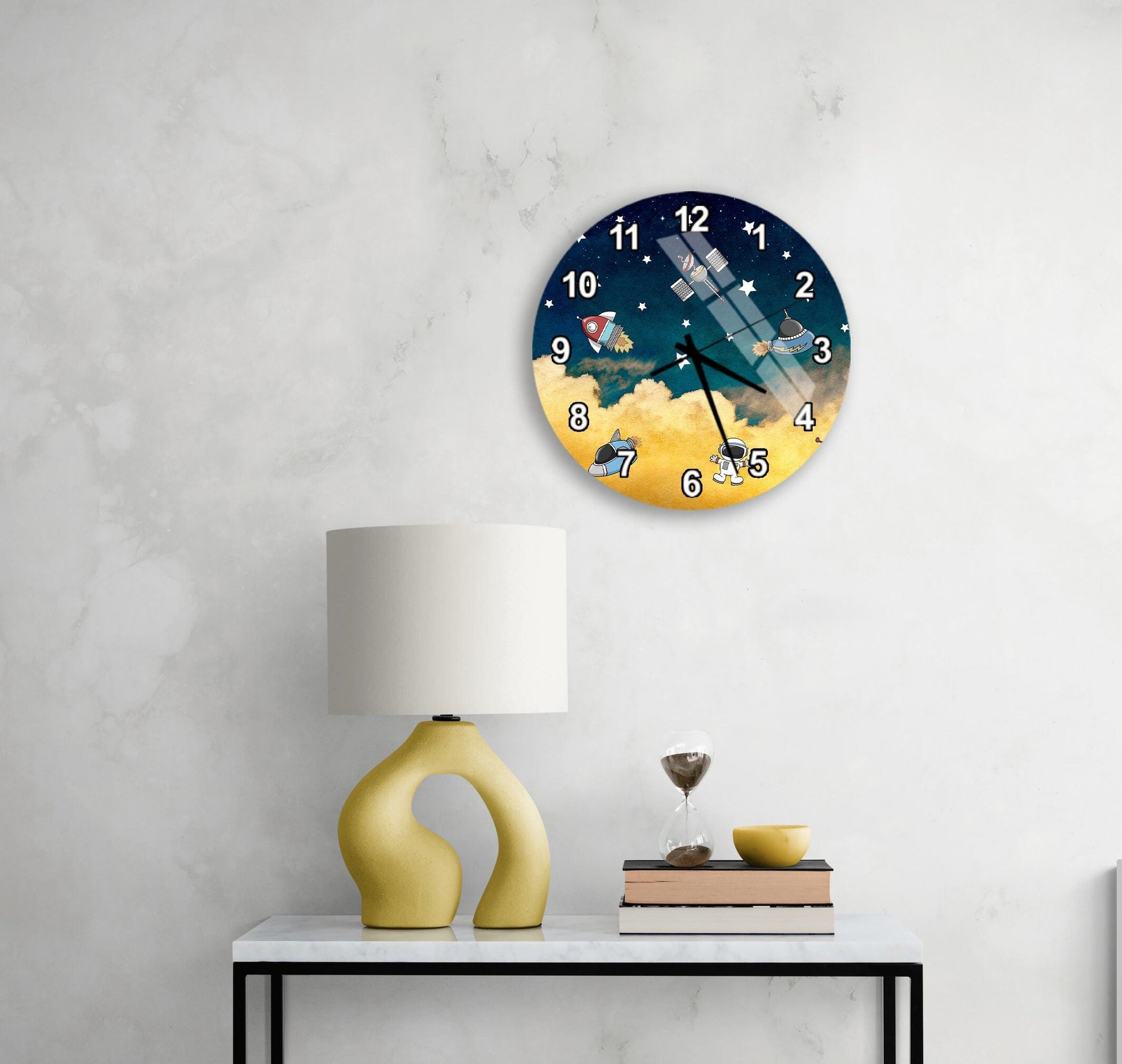 a round clock with a space theme, mounted on a wall above a white table with a yellow lamp and a stack of books.