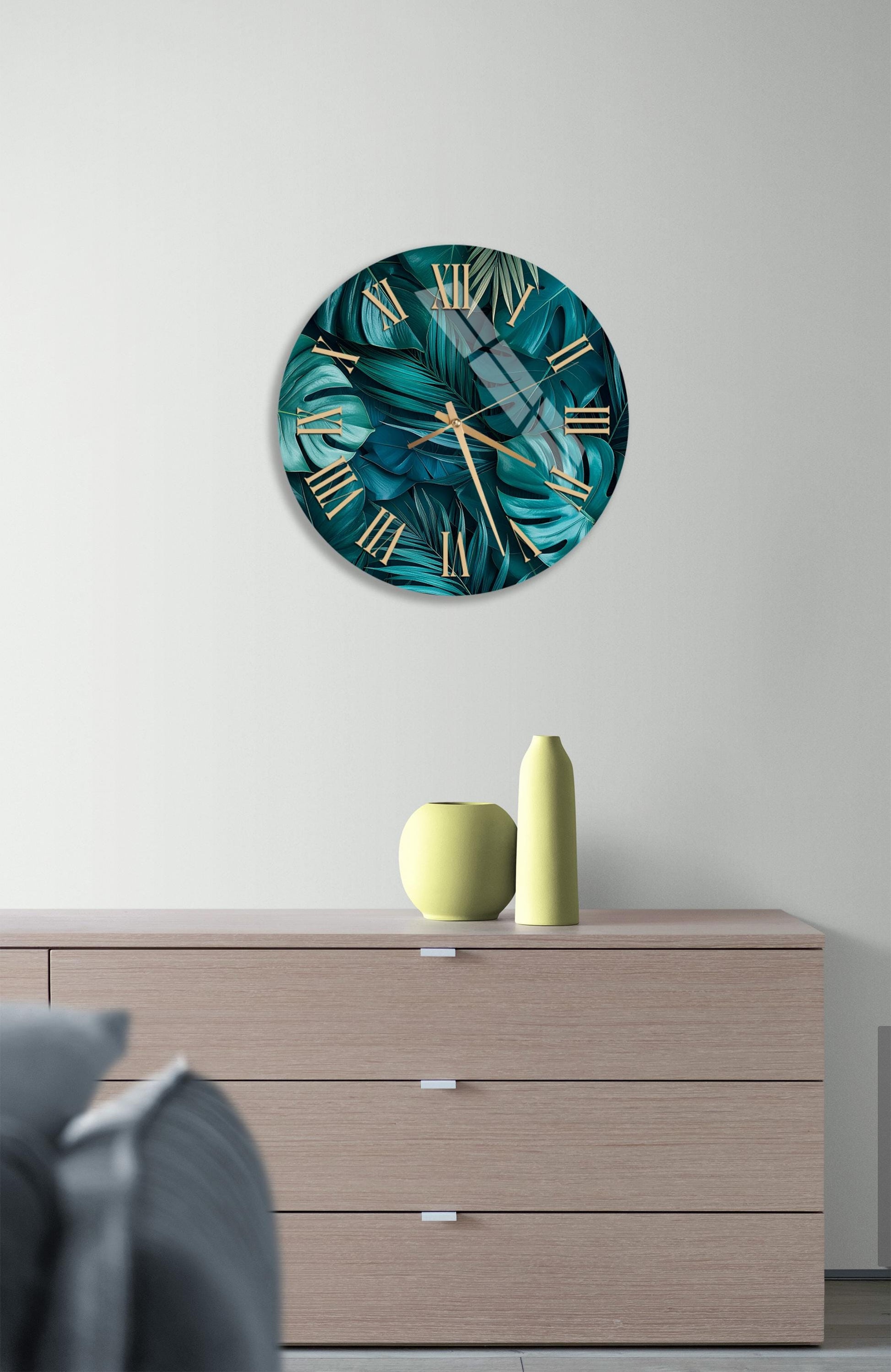 a modern living room with a large wall clock displaying a tropical leaf pattern in shades of green and blue. The clock is mounted above a wooden dresser with three drawers. On the dresser, there are two yellow vases and a small white vase.