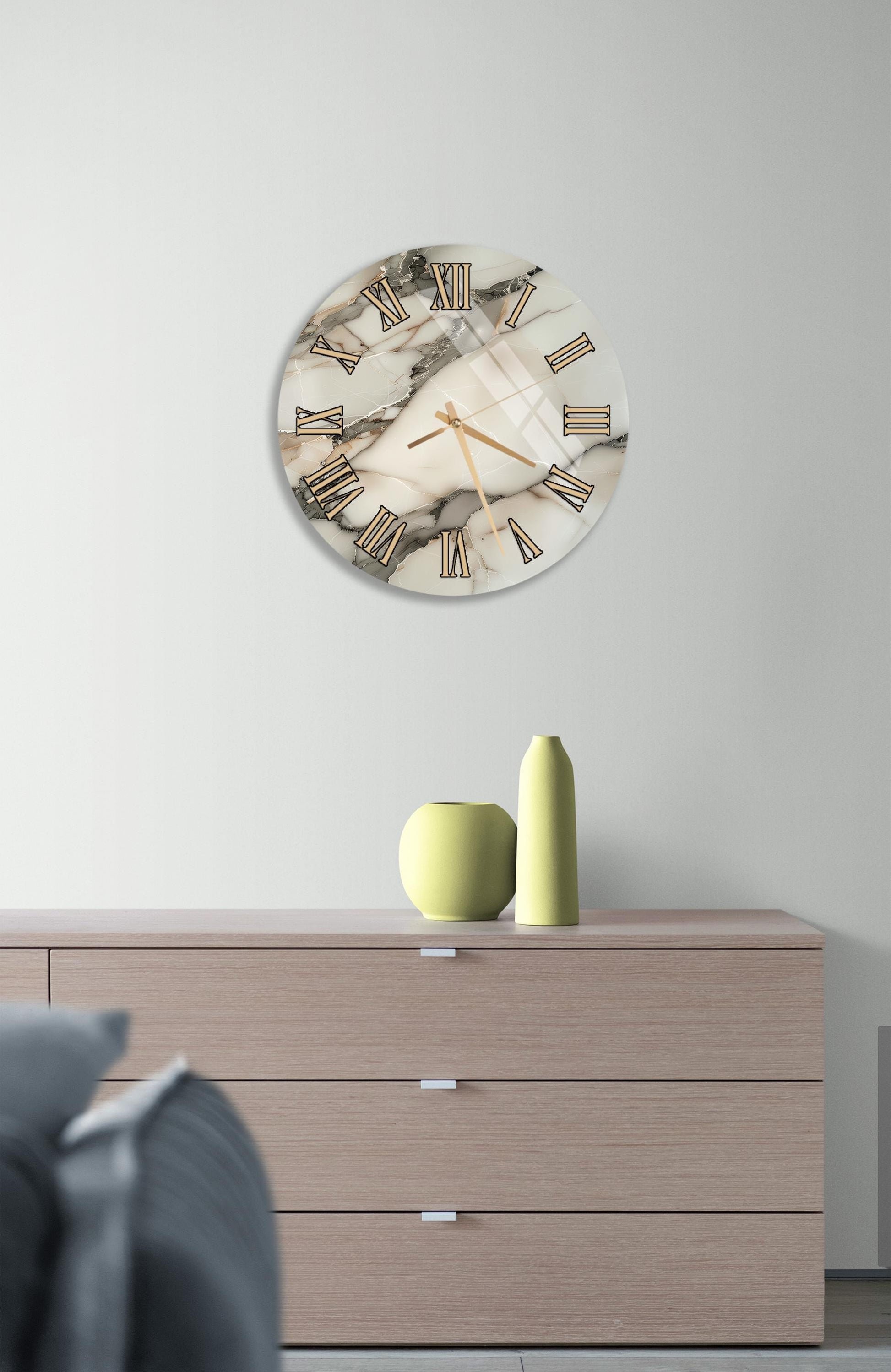 a modern living room with a round wall clock displaying Roman numerals, a wooden dresser with a vase and a small green sculpture, and a gray couch in the background.