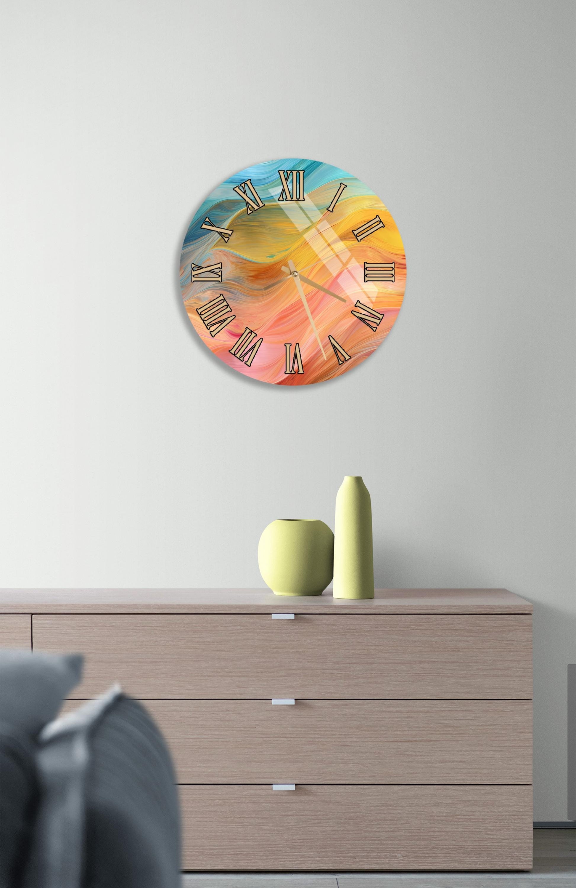 a wall clock with a colorful abstract design, mounted on a wall above a wooden dresser with a vase and a small sculpture on top.