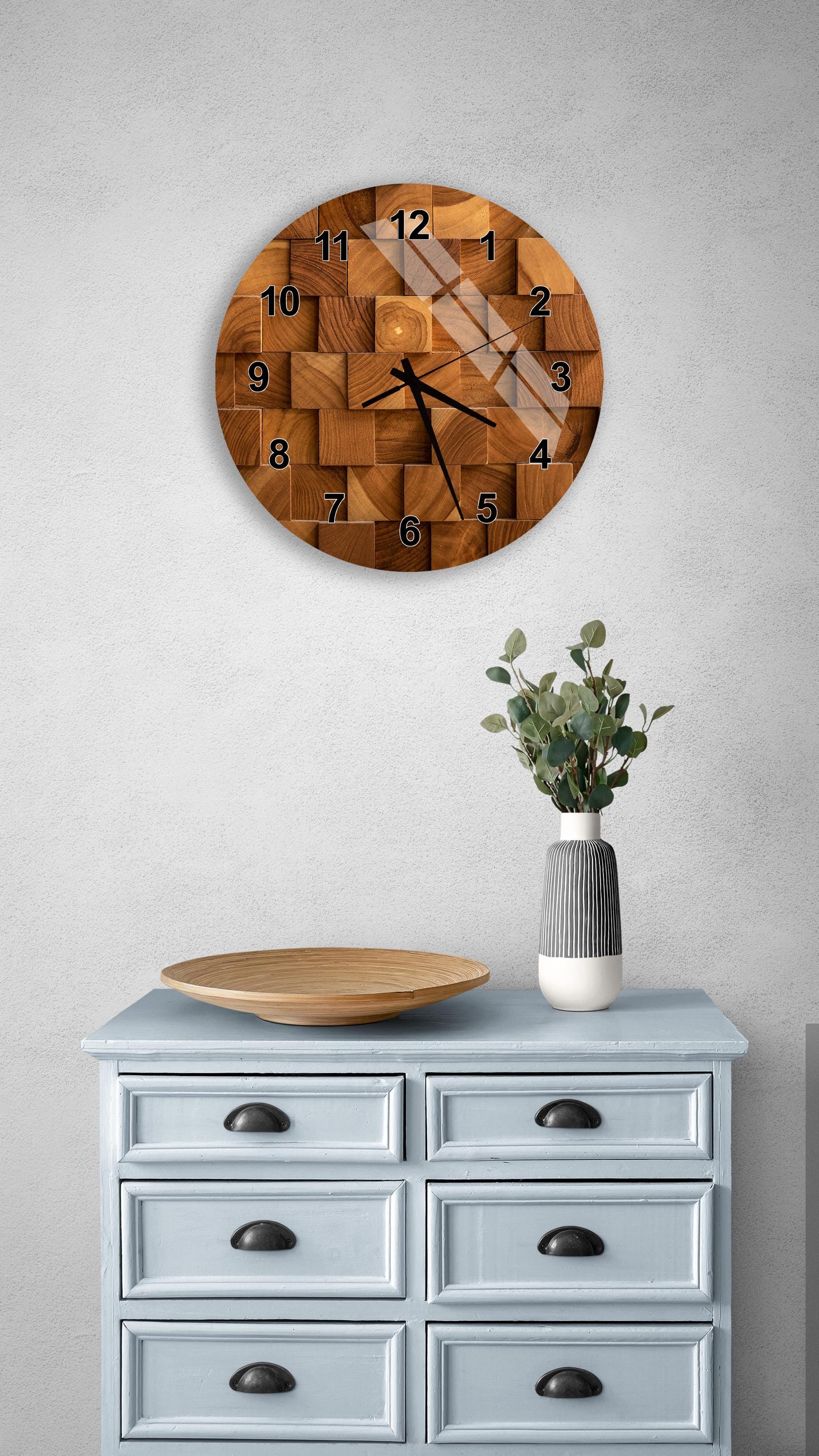 a wooden clock with a unique geometric design, mounted on a wall above a light blue dresser with six drawers. The clock has a wooden base and a wooden face with black numbers and hands. The dresser has a vase with greenery on it, and there is a wooden plate on top of it.
